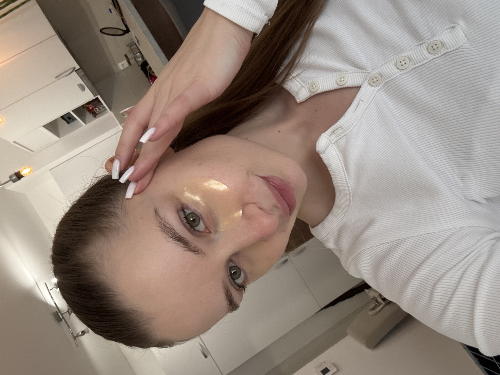 A woman with brown hair, green eyes, and a golden patch under her left eye is posing for a photo. Her left hand with white nails is positioned near her forehead. She is wearing a white, long-sleeved, button-up shirt. The background appears to be a kitchen with white cabinets. The location is likely indoors. The city of Budva, Montenegro cannot be determined from the image.