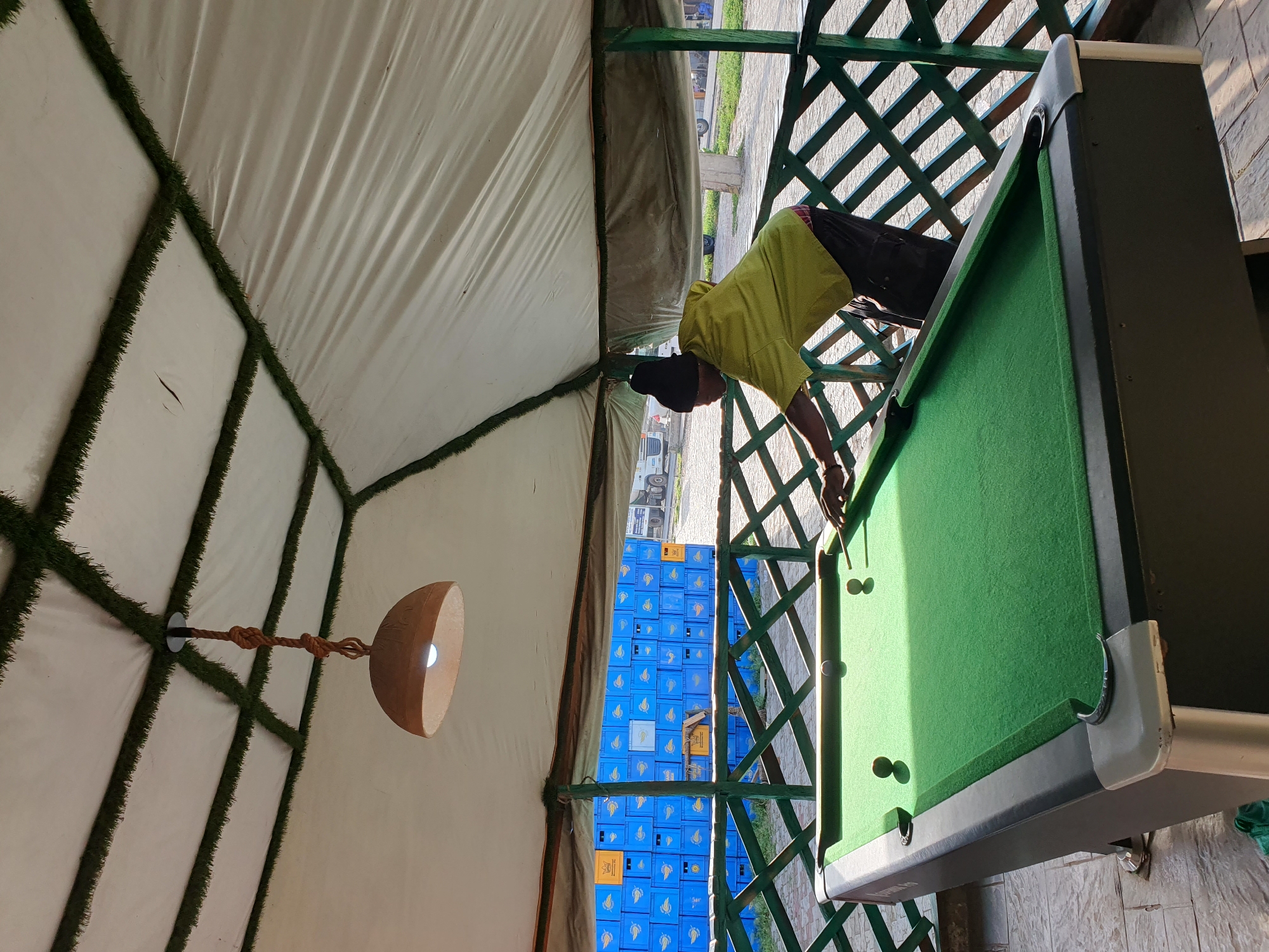 In Lokoja, Nigeria, a man wearing a green shirt and black beanie plays pool under a covered, open-air structure. The structure's ceiling is white tarp with green artificial turf along the edges of the structural supports. A hanging lamp with a brown shade illuminates the pool table. The pool table has a green felt surface and a few balls are visible. The man is in the process of taking a shot. In the background, stacks of blue crates are visible, and beyond that, trucks and a road suggest an outdoor, possibly commercial or transportation hub, setting. A green lattice fence surrounds the pool table area.