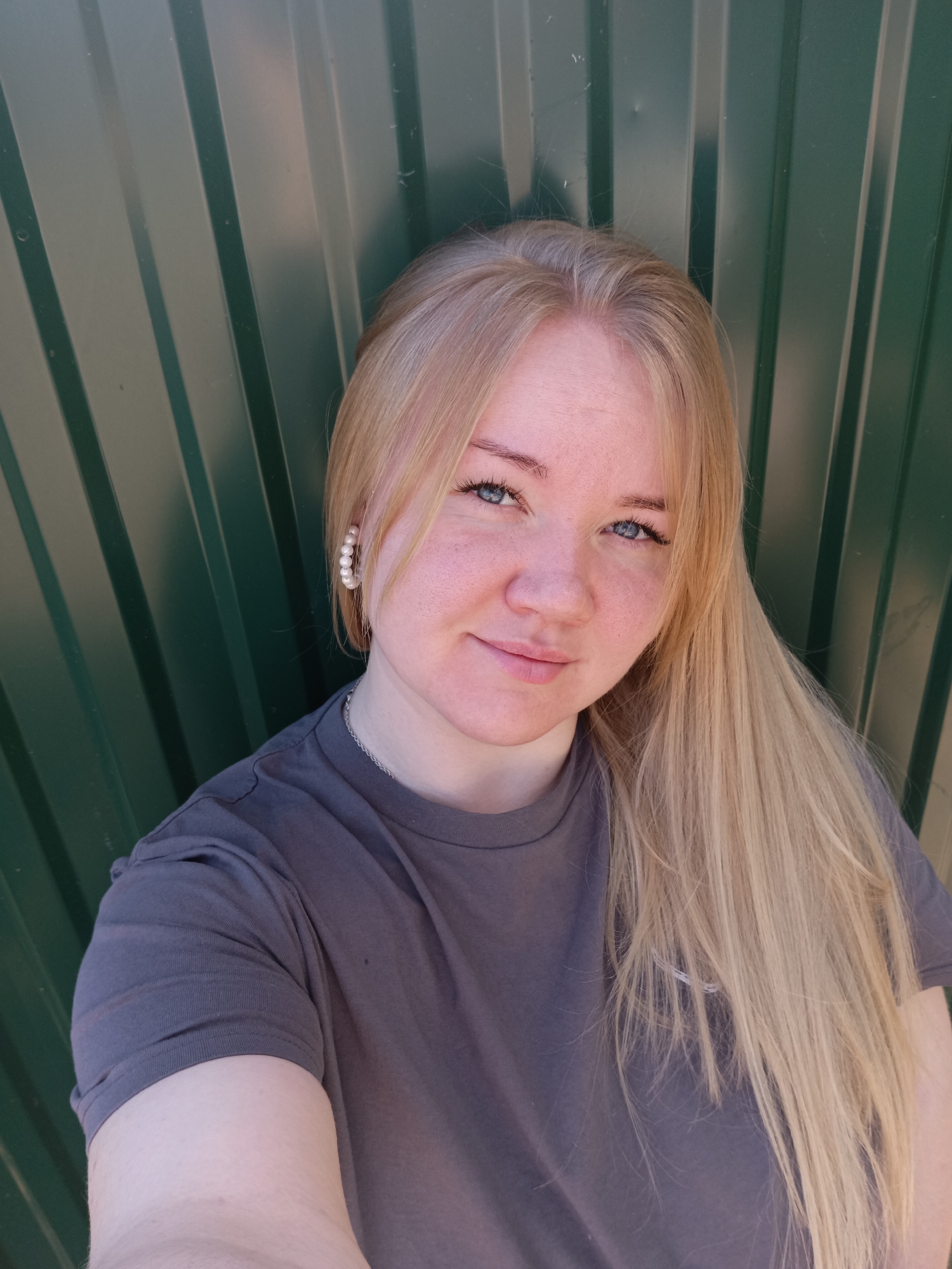 A light-skinned young woman with blonde hair and blue eyes poses for a selfie against a green corrugated metal wall. She wears a gray t-shirt and pearl hoop earrings. The location in the city of Фоки, Russia is not discernible from the image.