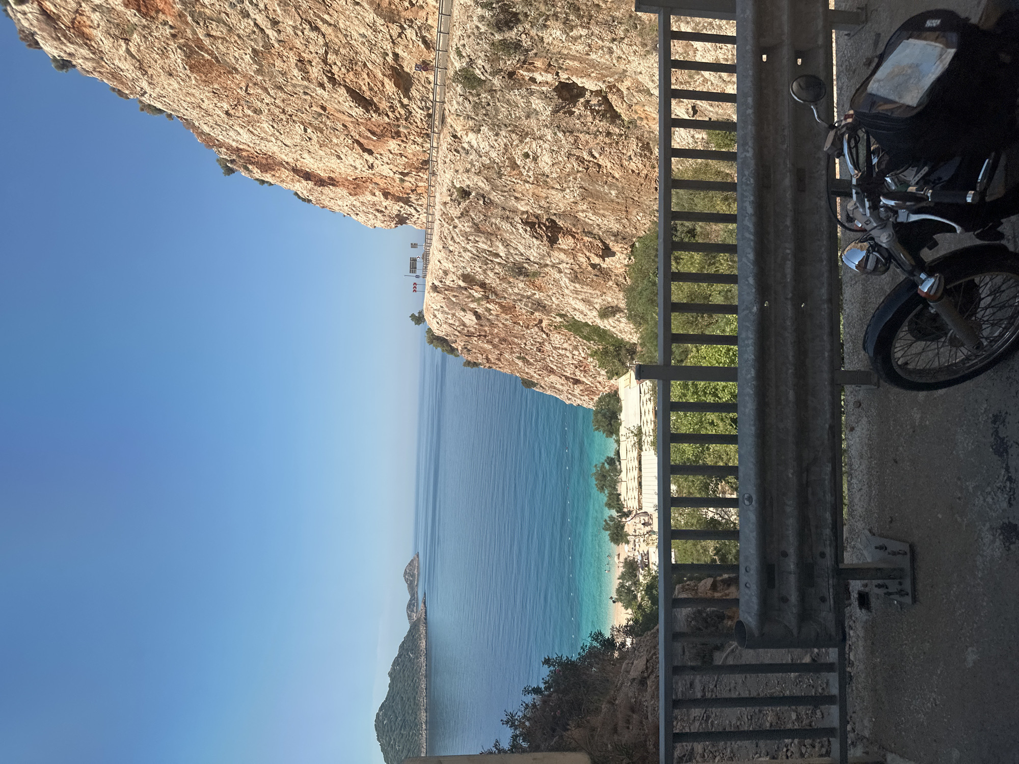This image captures a breathtaking coastal landscape, likely viewed from a road built into rugged cliffs. The scene is dominated by dramatic, sun-baked rock formations, a clear blue sky, and the stunning turquoise-to-deep-blue waters of the sea.
In the foreground, a sturdy metal guardrail runs vertically along the right side of the frame, suggesting a viewpoint from a cliffside road or overlook. Partially visible to the right of the guardrail is the front section of a black motorcycle, including its handlebars, rearview mirror, chrome detailing, and front wheel. A black tank bag is also noticeable on the motorcycle.
Beyond the immediate foreground, the view opens up to a serene bay or cove. A pristine sandy beach curves along the base of the cliffs, dotted with numerous white structures, likely sun loungers and umbrellas. Small figures of people can be seen enjoying the beach and the shallow, clear waters. Several small boats or buoys are visible in the sea near the shore. Distant landmasses or islands appear as dark silhouettes on the horizon, blending with the deeper blue of the sea.
Towering cliffs rise steeply from the water on the left side of the image, showcasing their textured, reddish-brown rock faces. Further up these cliffs, a winding road with its own guardrail can be seen, leading to a distant, higher vantage point. Small patches of green vegetation cling to the rocky slopes.
The image depicts a bright, clear day, likely late morning or early afternoon, with ample sunlight illuminating the landscape. The weather appears perfect for outdoor activities. The overall setting strongly aligns with coastal regions like Kaş, Turkey, known for their stunning coves and rugged beauty. No discernible text is visible within the image.