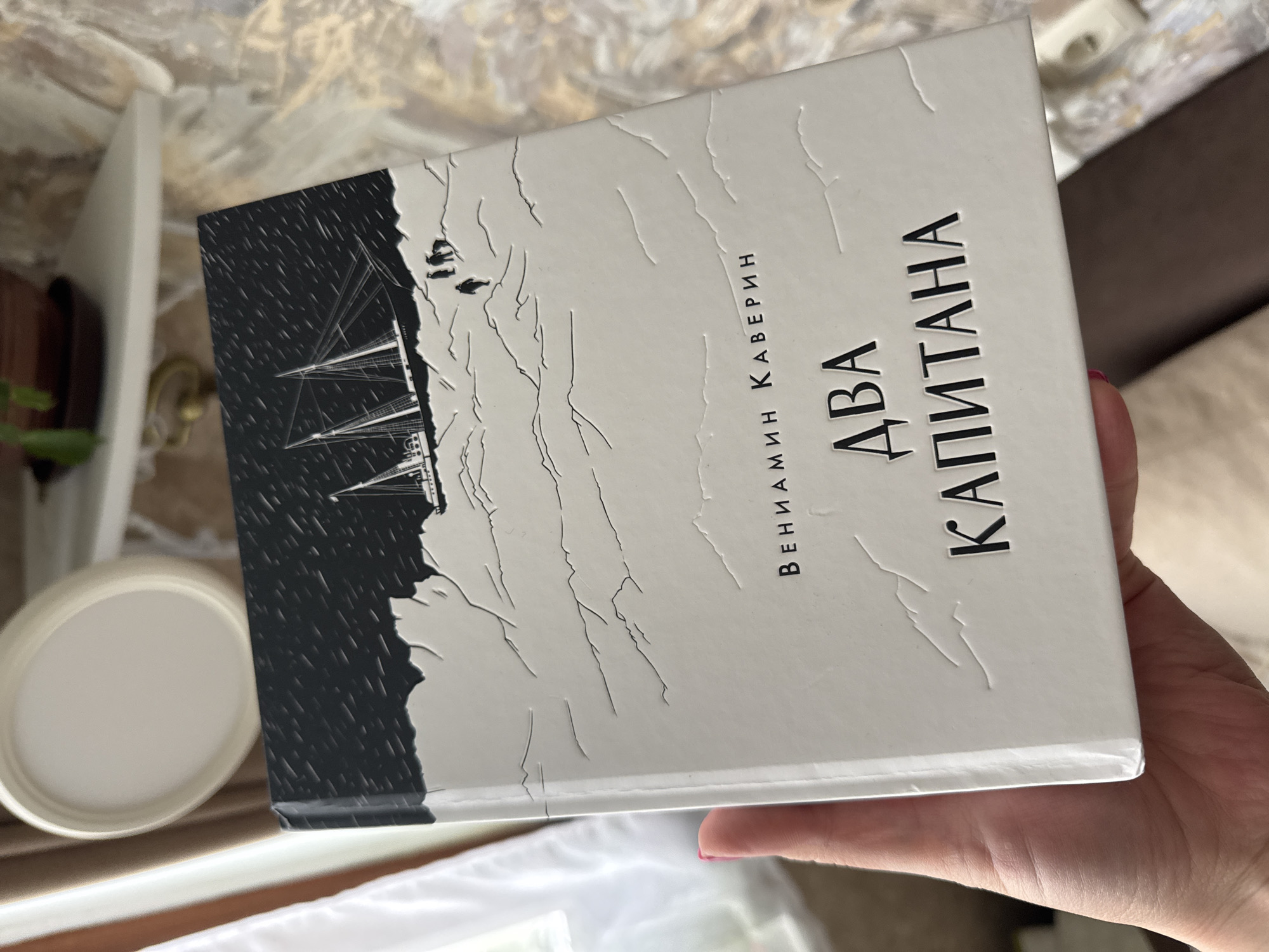A person's hand holds a book titled "ДВА КАПИТАНА" (Two Captains) by "ВЕНИАМИН КАВЕРИН" (Veniamin Kaverin). The book cover is predominantly white with a prominent black section on the left. This black area depicts a multi-masted sailing ship, possibly a schooner or barque, amidst what appears to be falling rain or snow. The white portion of the cover features textured, raised lines that resemble cracks in ice or a rugged, snowy landscape. Two small, dark, silhouetted figures, possibly people, are visible on the white part of the cover, positioned near the black section. The hand holding the book has a pink-painted fingernail visible on the thumb. The background is an out-of-focus indoor setting with warm, neutral tones, showing glimpses of a wooden surface, a white round object (possibly a plate or coaster), a green plant, and light-colored fabric. The book is held slightly tilted, presenting its cover prominently. The overall impression is quiet and domestic.