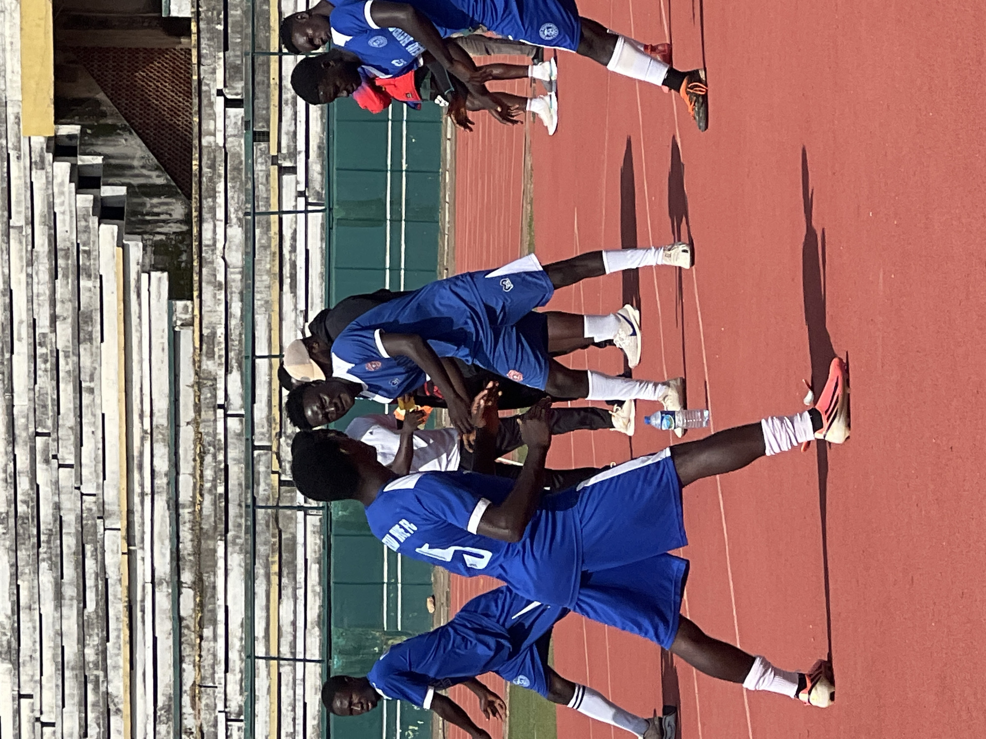 In New Stadium, Nigeria, a group of young men in blue soccer uniforms are on a red track. The stadium seating, mostly empty and weathered, is visible in the background. Two men in the foreground are facing each other, their arms outstretched in what appears to be a drill or exercise. One man has the number "5" on his jersey. Other players are scattered around the track, some observing, others engaged in their own activities. A water bottle sits on the track near the foreground. The lighting suggests it is daytime, with the sun casting distinct shadows.