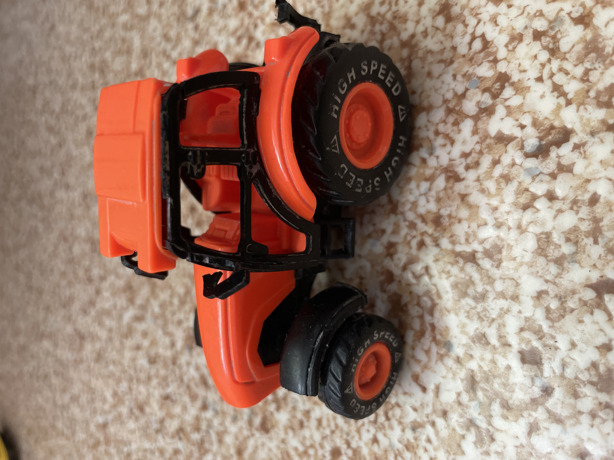 An orange plastic toy tractor is positioned on a speckled, light brown surface. The tractor has a black roll cage and black tires. The front tires are smaller than the rear tires. The words "HIGH SPEED" are visible on the sidewall of the rear tire, along with a triangular symbol. The tractor is facing towards the left of the frame. The background is out of focus and does not provide any specific environmental details. There is no information about the city of Laishevo, Russia, present in the image.