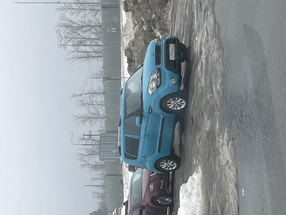 This image depicts an outdoor scene in Sholokhovo, Russia, under an overcast sky. The primary subject is a light blue (cyan) first-generation Kia Soul, bearing Russian license plate `O254EX71`. The vehicle is parked on a wet, dark asphalt or tarmac surface, alongside a partial accumulation of dirty snow and slush. To the left and slightly behind the Kia, a dark red sedan, possibly a Skoda Octavia, is partially visible. To the right of the Kia, a large mound of dirty snow mixed with earth and debris is present. The immediate foreground shows patches of melting snow and standing water on the dark pavement, with a clear plastic bottle lying horizontally. In the background, a light grey sound barrier wall extends horizontally, behind which a line of bare deciduous trees, utility poles, and streetlights are visible against a uniformly pale sky. No individuals are present in the frame.