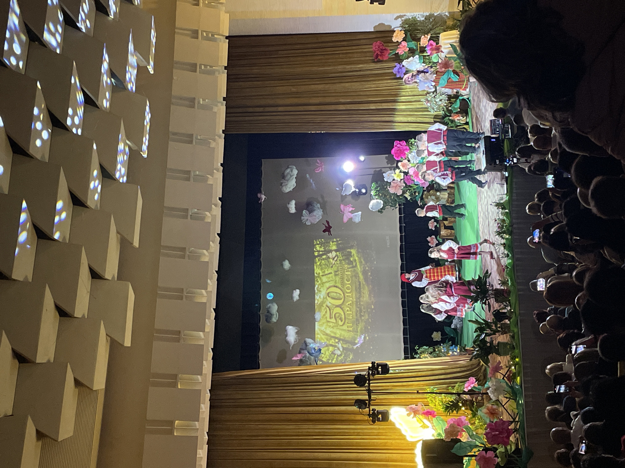 This image captures an indoor stage performance from an audience perspective, looking towards a brightly lit stage. On the left, a textured wall features numerous geometric, block-like structures, some of which are illuminated with colorful, speckled light patterns.
The stage is framed by rich, golden-brown pleated curtains. It is elaborately decorated with a vibrant array of large, artificial flowers in shades of pink, red, white, and purple, interspersed with green foliage. Spotlights illuminate these decorations and the performers on stage.
Several performers, primarily children and young adults, are visible on stage. They are dressed in traditional folk costumes, predominantly featuring red, white, and black colors with embroidered patterns. The costumes suggest a cultural or folk dance performance.
Behind the performers, a large projection screen displays a visual backdrop. The screen shows a green, possibly forest-like, scene with abstract white shapes resembling clouds or stylized flowers, and colorful butterflies. Prominently displayed on the screen is a large number "50" and Cyrillic text, including "РАДОСТ" (meaning joy or happiness). The word "FREEZE" is also visible in the top right corner of the projected area.
On the far right of the image, a large audience is visible, mostly as dark silhouettes in the dimly lit seating area. Many audience members are holding up smartphones, capturing the performance. The event appears to be an evening celebration, possibly an anniversary (indicated by the "50"), taking place in Zlatograd, Bulgaria, given the cultural context of the costumes and location hint.