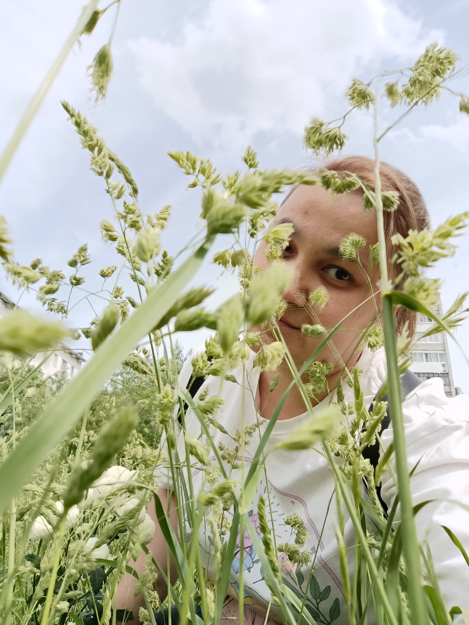 The image is a close-up shot of a person, likely a young adult, standing amidst tall grass with seed heads. The grass obscures parts of the person's face and body, offering a partial view of their face. They have fair skin, light brown hair and are looking towards the camera with a slight smile.
The person wears a white T-shirt with a faint floral pattern. A backpack strap is visible over their shoulder. Behind the grass and the person is a building that looks like an apartment block, suggesting an urban location. The sky is visible above, partially obscured by clouds.
While the prompt mentions Chaikovsky, Russia, there are no distinctive landmarks in the image to confirm the location.