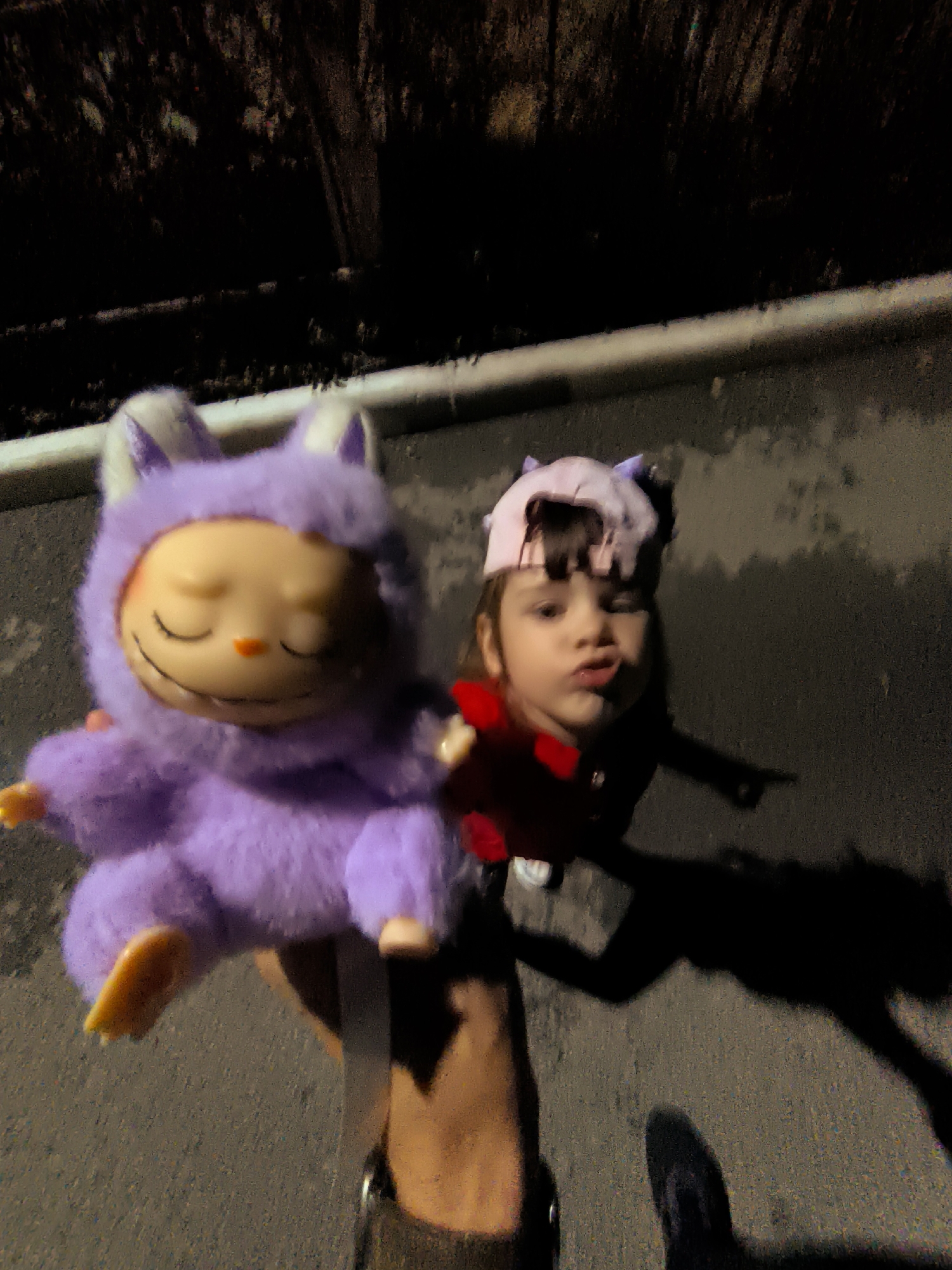 In an outdoor setting, a person is holding up a large, fuzzy, lavender-colored plush toy with white-tipped ears. The toy has closed eyes and an orange nose. To the right of the toy, a young child is visible, wearing a pink and purple hat and a red shirt. The child is looking towards the camera with pursed lips. The ground appears to be asphalt. A white pipe runs horizontally across the mid-ground, and in the background, there is a dark, textured surface, possibly foliage or tree bark. The lighting suggests it is nighttime or a dimly lit area. The scene is a snapshot of a playful moment outdoors, with the child and the toy being the primary focus.