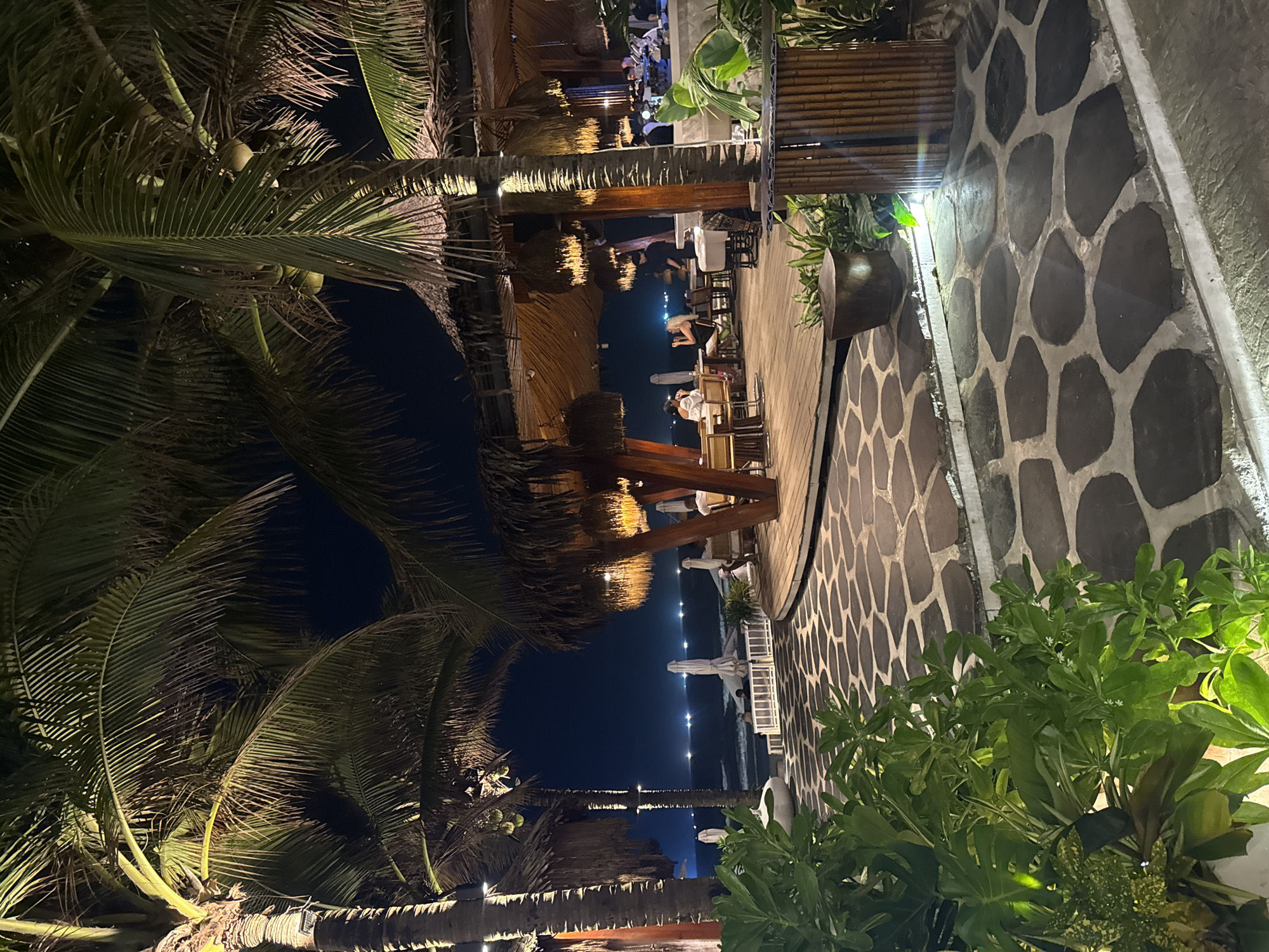 The image shows a nighttime scene in an outdoor restaurant in an unknown location. The restaurant has a wooden deck with tables and chairs under a thatched roof. There are people dining at the tables. Palm trees surround the area, and their leaves are visible in the foreground and background. A wall made of irregularly shaped stones is to the right, adorned with foliage. Beyond the restaurant, lights are visible along the water's edge.