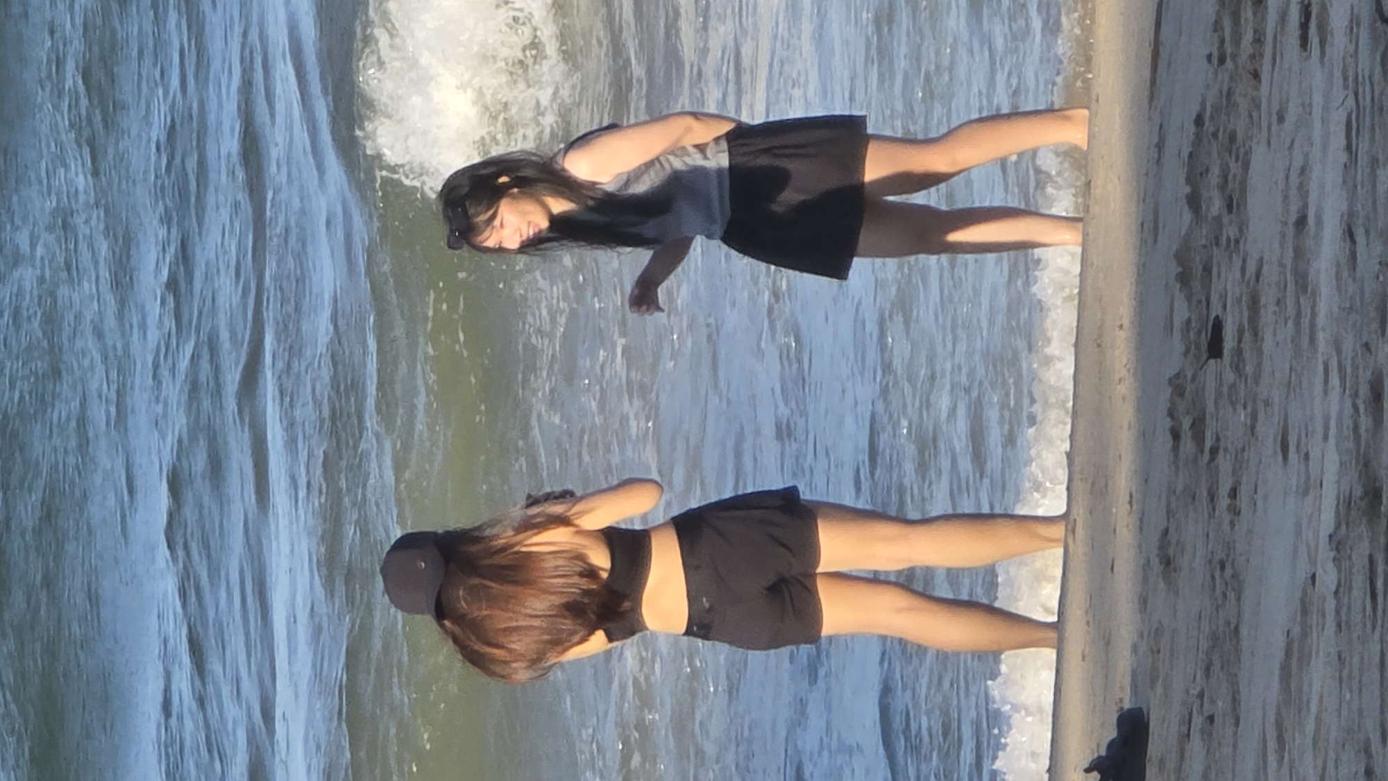 Two women stand on the beach at Nha Trang Ward, Vietnam. One woman wears a gray tank top and a black skirt; the other woman wears a black sports bra, black shorts, and a baseball cap. Both women stand at the water's edge. The beach sand is tan and wet. The sea water is foamy.