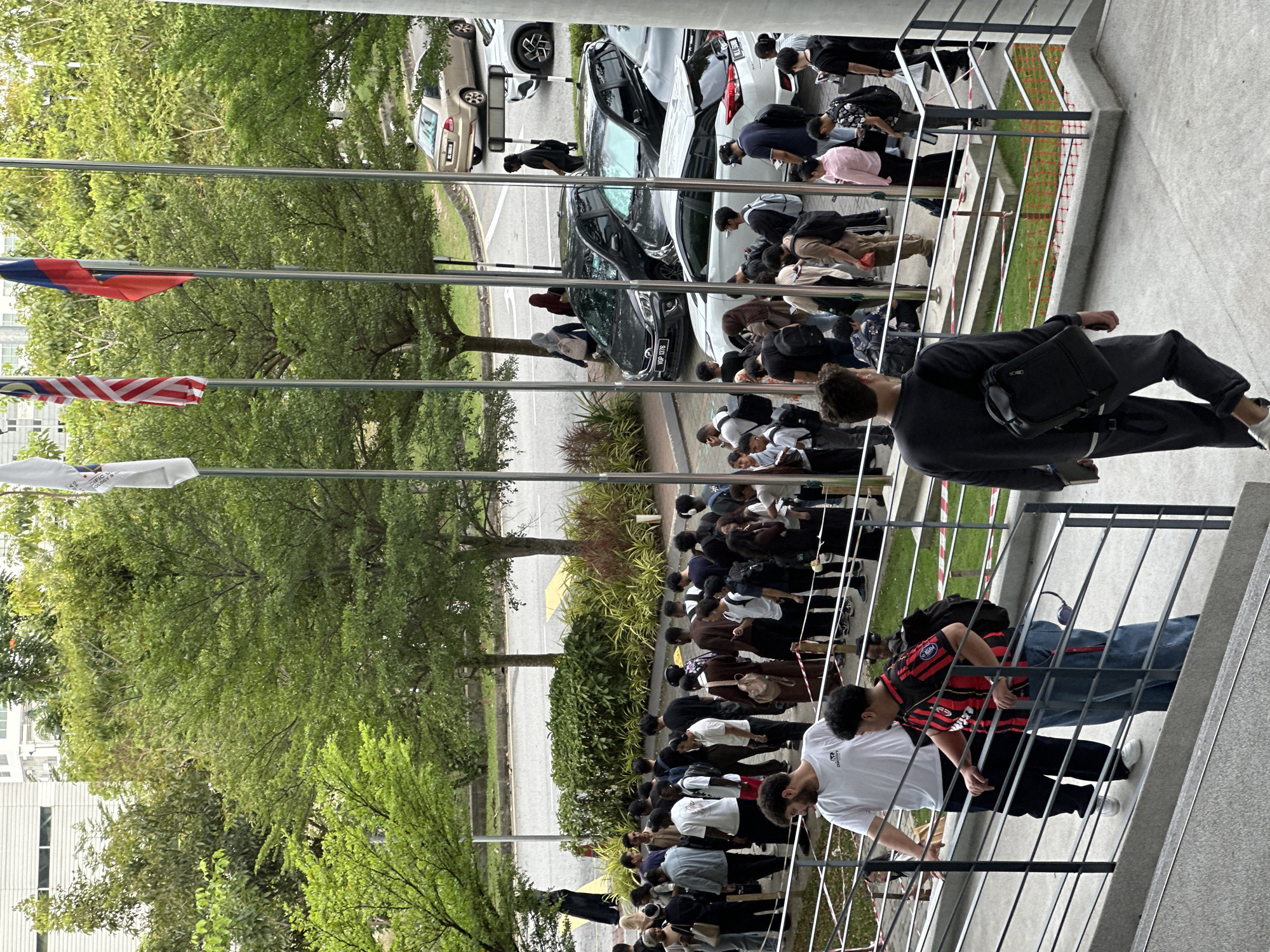 The image shows a large group of people standing in a line outside a building. They appear to be of various ages and ethnicities. The people in the line are wearing a variety of casual clothing. Several cars are parked on the street nearby. Flags are visible. The location is likely Kuala Lumpur, Malaysia, as flags of Malaysia are visible.