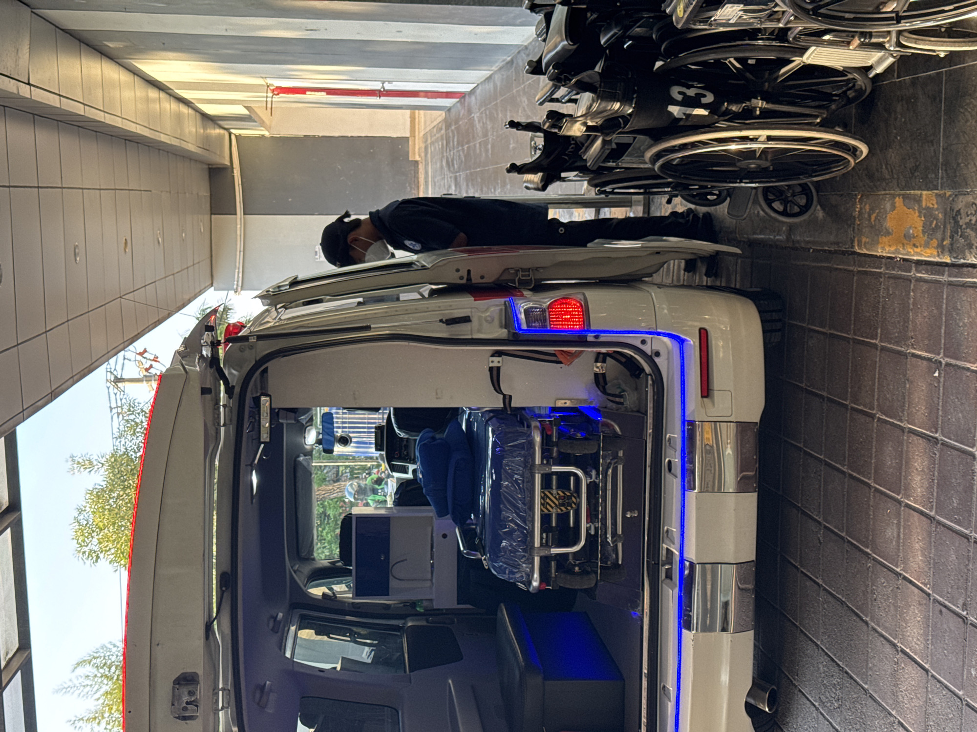 An ambulance van is parked with its rear doors open, revealing a stretcher covered in blue plastic. Inside the van, a blue bench seat and a sink are visible. Blue LED lights illuminate the rear interior of the van and the bumper. To the right of the van, a person wearing a dark uniform, a black cap, and a white face mask is standing near several stacked wheelchairs. The scene takes place on a paved walkway with tiled flooring, possibly near a building entrance in Kuta, Indonesia, as suggested by the context of the image. The lighting indicates it is daytime, with sunlight casting shadows.