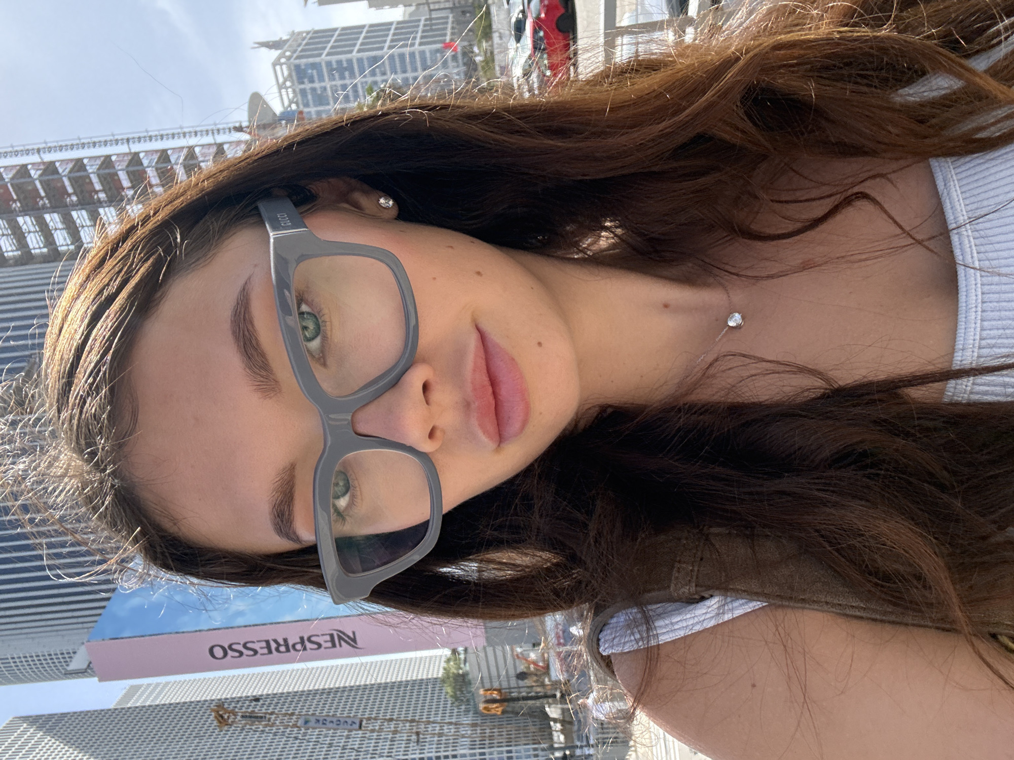 A woman with long brown hair and gray-framed glasses is in an outdoor setting. She wears a white top and a delicate silver necklace with a single round pendant. Her left ear is visible, adorned with a stud earring. The background features a cityscape, including tall modern buildings and a pink and blue Nespresso advertisement. The location is likely Tel Aviv, Israel, given the architectural style of the buildings. The overall lighting suggests a bright, sunny day.
