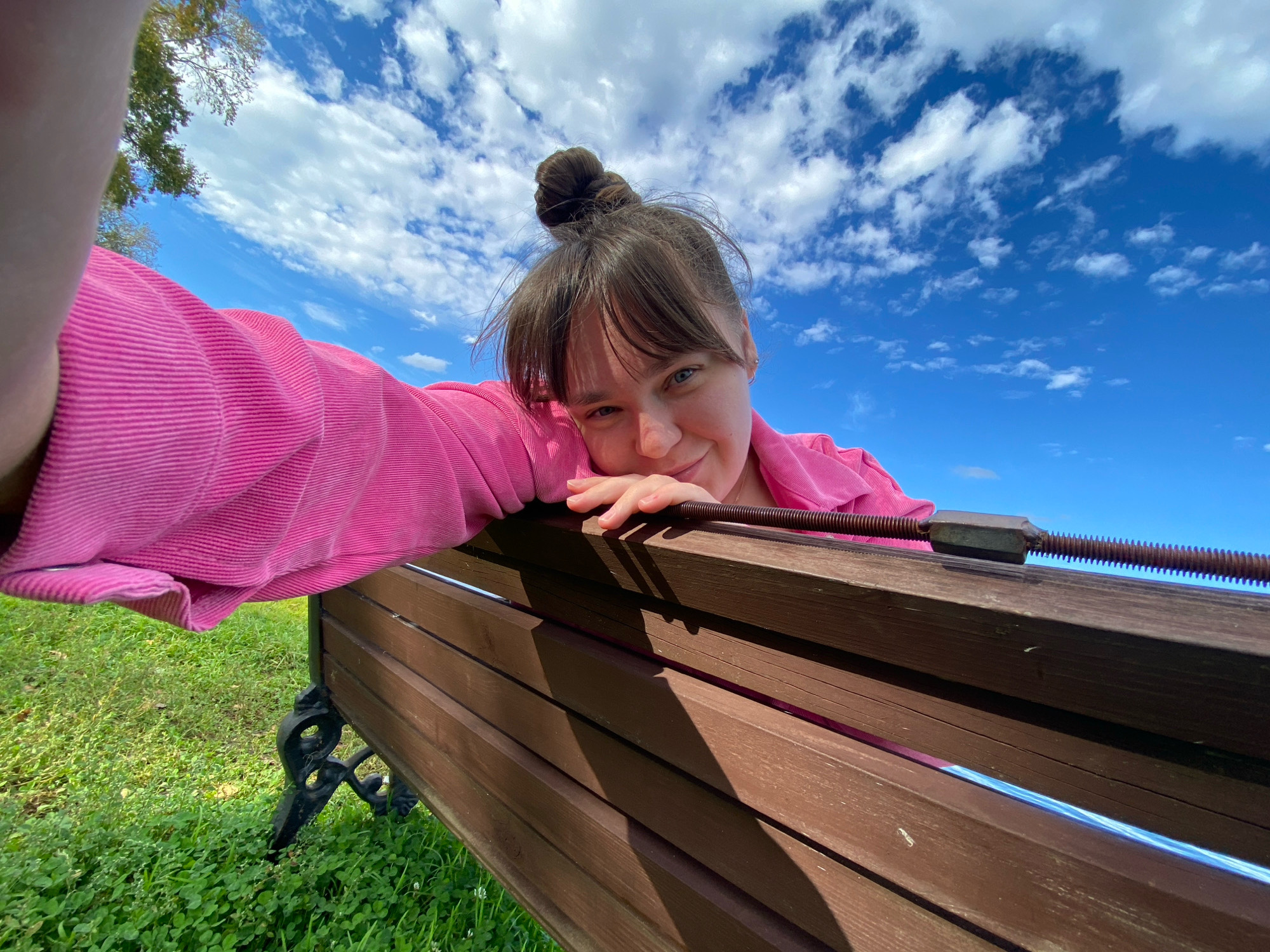 A low-angle, wide shot shows a young woman with her hair in a bun, leaning over a wooden park bench. She is wearing a vibrant pink, corduroy shirt. The bench has dark metal legs and is surrounded by green grass and clover. In the background, a bright blue sky is filled with fluffy white clouds, and a tree with green leaves is visible on the left. The woman is smiling and looking at the camera, suggesting a happy and relaxed mood. The lighting indicates it's daytime, likely on a sunny day with some clouds. The image appears to be a selfie taken outdoors.