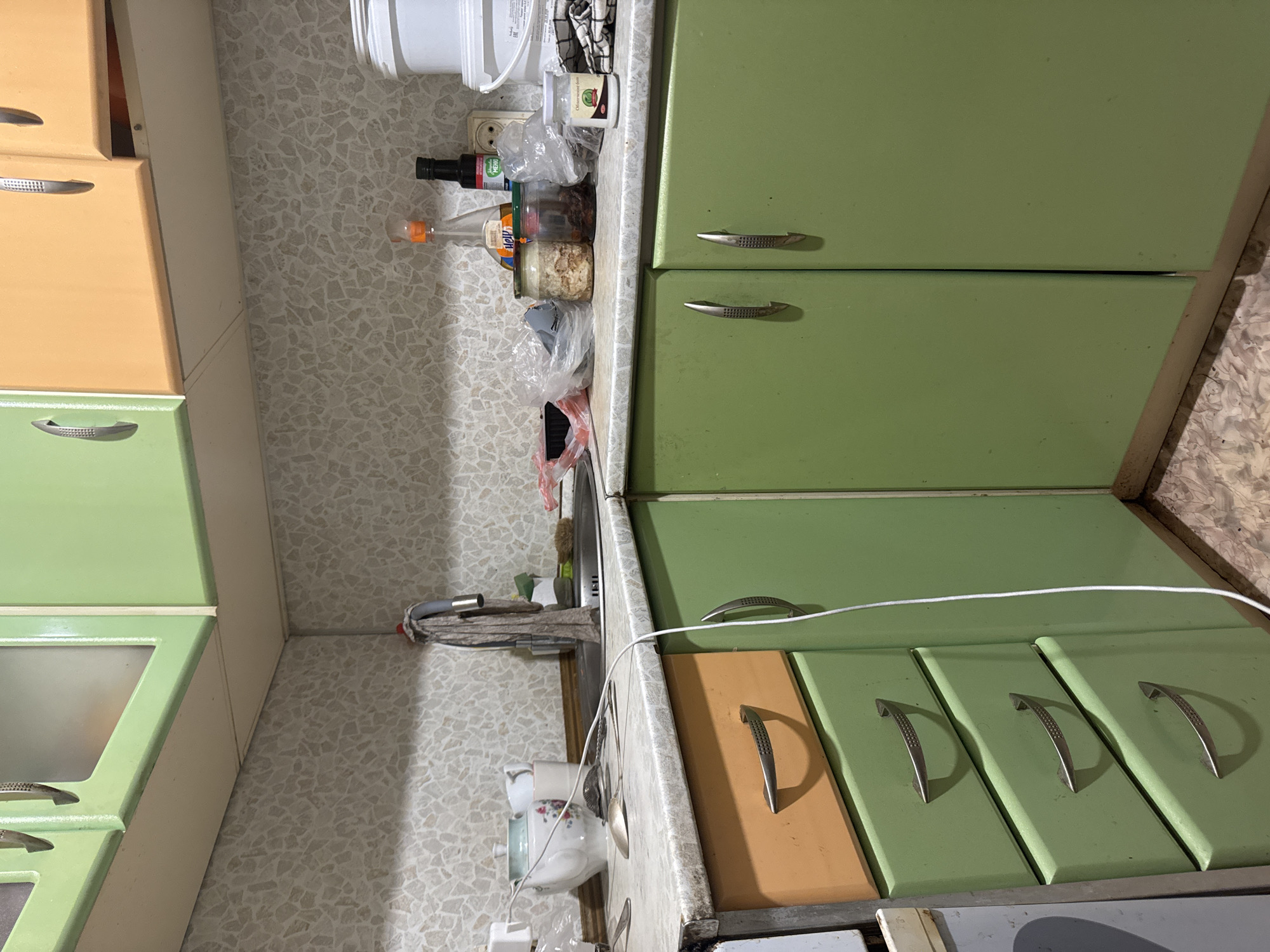 This image depicts a cluttered kitchen counter and cabinets in a residential setting. The cabinets are painted in two shades: a pale orange on the upper section and a light green on the lower section, with silver handles. The countertop is a speckled grey laminate.
On the counter, to the left, a white teapot with a floral design and a white cup are visible. A sink with a faucet and a dishtowel hanging from it is located towards the back. Various items are scattered around the sink area, including plastic bags containing food items, two glass jars with contents, a bottle of soy sauce, a bottle of cooking oil, and a small jar with a label that appears to be in Cyrillic. To the far right, a large white bucket or container is present. A white electrical cord runs diagonally across the scene.
The backsplash is a beige wallpaper with a mosaic-like pattern of irregular grey and white tiles. The floor is tiled with a light-colored material featuring a speckled, irregular pattern.
The image does not provide any information that can definitively place it in the city of Демихово, Russia, or any other specific location. The scene appears to be a typical, lived-in kitchen environment. There are no people visible in the image, and no specific actions or interactions are depicted. The lighting is somewhat dim, suggesting it might be indoors with artificial lighting.