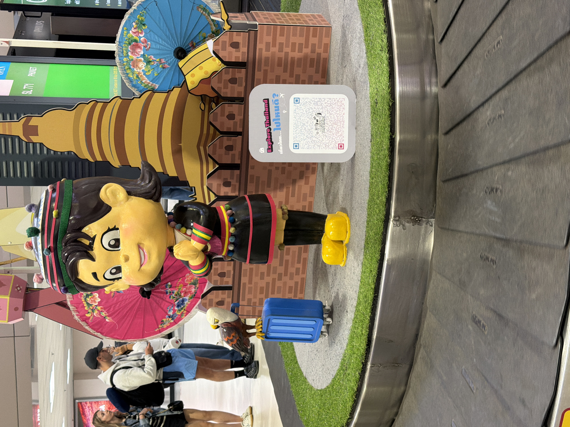 This image depicts an indoor scene at an airport baggage claim, likely in Bangkok, Thailand, featuring a vibrant cultural display next to a baggage carousel. The central focus is a large, cheerful statue of a character, possibly a child or cartoon figure, dressed in traditional Thai-like attire with a distinctive multi-colored headband and yellow shoes.
The display is set on a raised platform covered with artificial grass and includes several other elements of Thai culture. Behind the main character, there's a golden, tiered stupa-like structure (chedi) and two ornate, floral-patterned Thai parasols, one blue and one pink. A brick-patterned backdrop forms part of the installation. A small blue suitcase with wheels sits near the statue's feet, accompanied by a small bird statue, possibly a Garuda, wearing a travel hat and holding the suitcase handle.
A prominent white sign on the display platform features two QR codes. Text on the sign reads "Explore Thailand" and the Thai phrase "เที่ยวไทย...ไปใหนดี?" which translates to "Travel Thailand... where to go?". Within one of the QR code designs, the words "LOVE TRAVELING" are also visible.
In the background, two individuals, likely passengers, stand near the display. A man wearing a cap, white hoodie, and blue shorts is looking at his phone. Next to him, a woman with long hair is partially visible. Further back, a digital display board shows flight information, including "SL771 PHUKET" and the word "OKAY". The "PHILIPS" logo is also seen on a panel. The dark grey, textured baggage carousel occupies the right side of the frame. The scene portrays a welcoming and culturally themed airport environment.