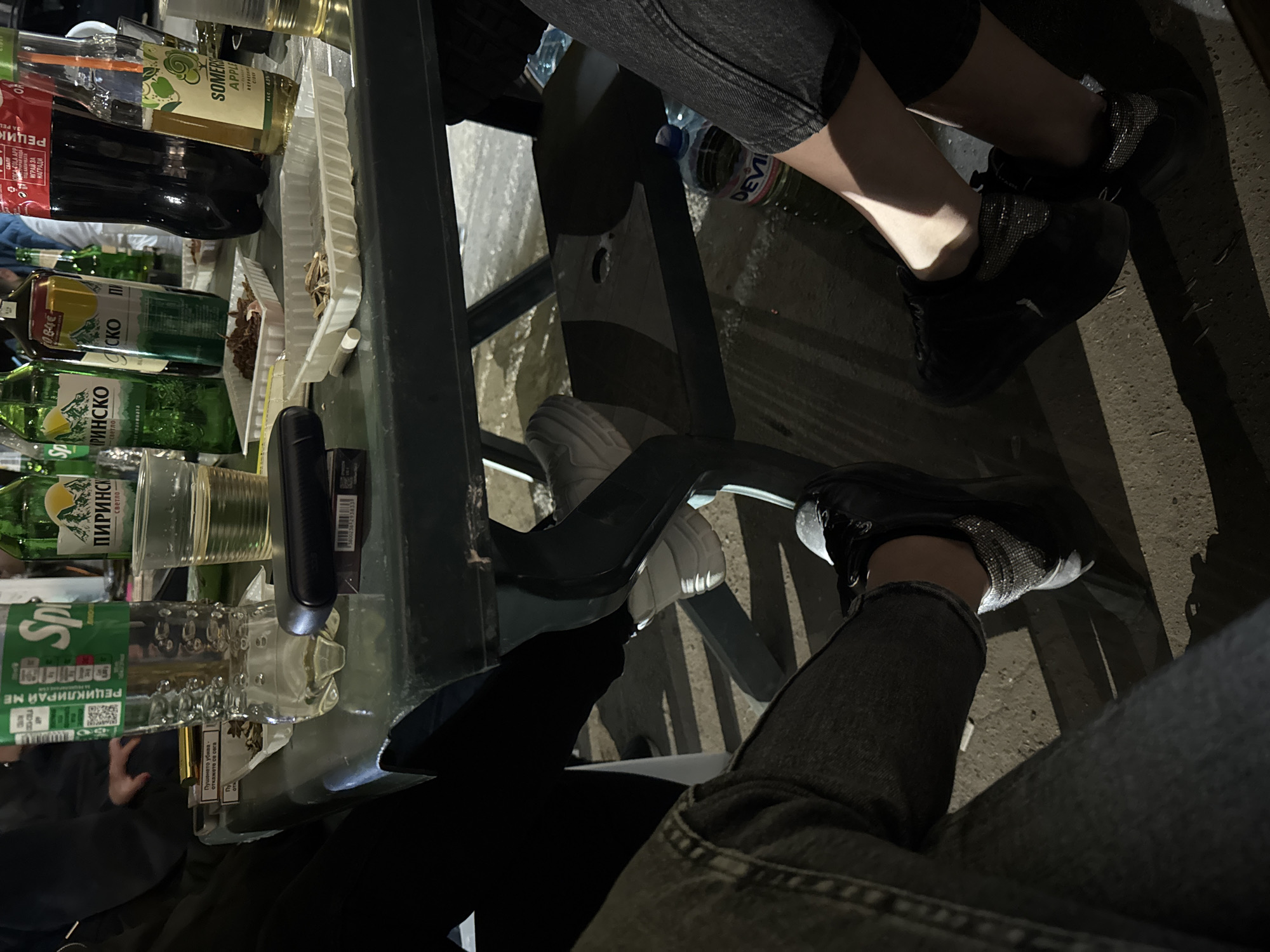 The image shows a table laden with various beverages, including bottles of Sprite, Pirinsko beer, Somersby cider, and Coca-Cola. A pack of cigarettes is also present. Two people are sitting at the table, their legs visible. Both individuals are wearing jeans and dark-colored sneakers, one pair with sequined detailing. The location is assumed to be in Topolovets, Bulgaria. The scene depicts a social gathering where drinks and cigarettes are consumed.