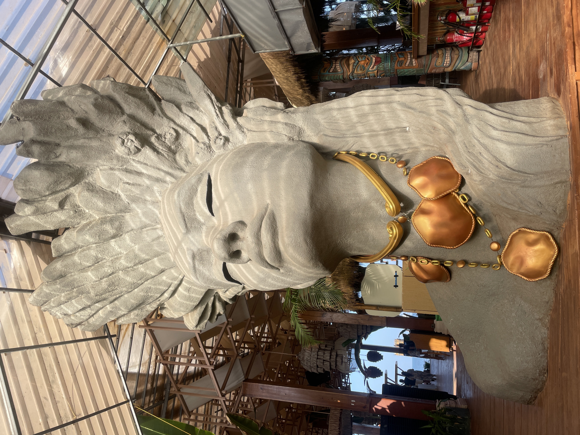 A large stone statue of a woman with closed eyes, adorned with a gold necklace, dominates the scene. The statue is indoors, beneath a thatched roof and a modern metal and glass structure. To the right of the statue, a wooden wall and decorative carvings are visible. Through an opening beneath the statue, an outdoor scene with people and structures is visible in the distance, suggesting the location is Chopdem, India.