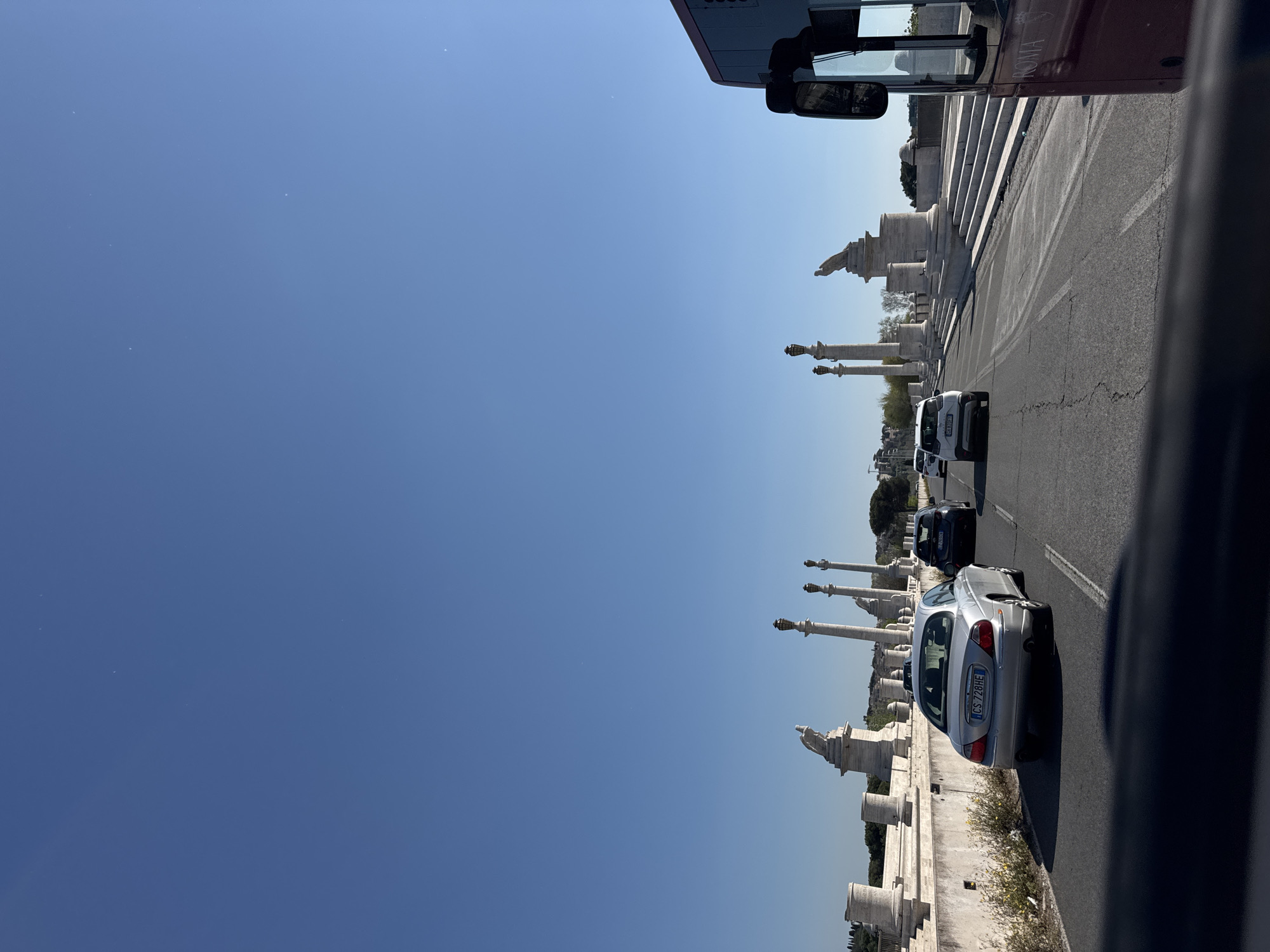 The image, taken from a moving vehicle, depicts a view of the Ponte Vittorio Emanuele II in Rome, Italy. A line of cars is traveling along the bridge, with the statues and architectural details lining the parapet visible on the right. The sky is clear and blue. Part of the bus is visible at the top of the image.