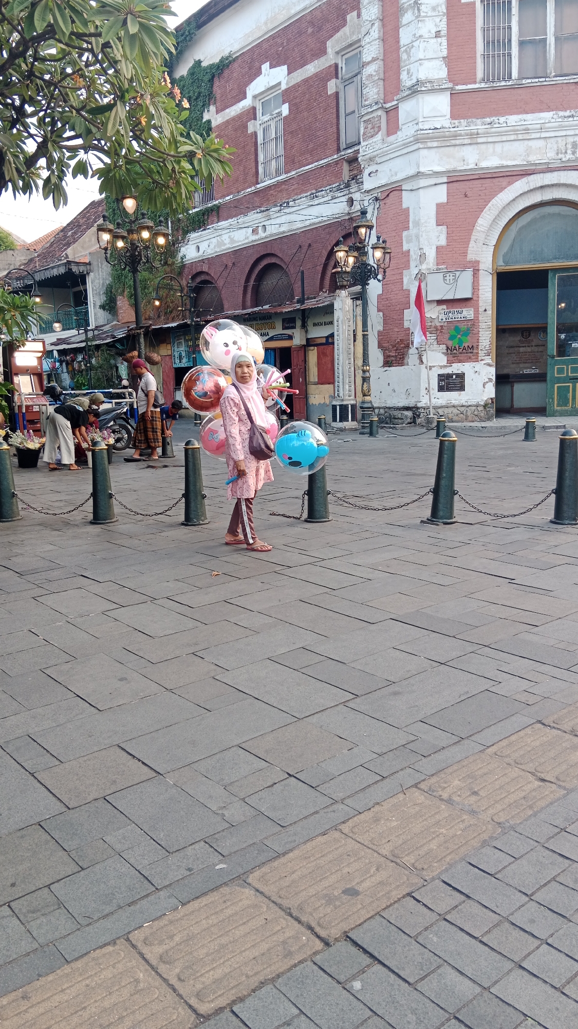 A woman wearing a hijab and a pink outfit stands on a paved plaza in front of a red-brick building with white trim and arched windows. She is holding a bouquet of colorful balloons, some of which are clear and contain cartoon characters. The woman is facing the camera, with a slight smile. In the background, there are several other people, some of whom are interacting with a motorcycle. The plaza is lined with bollards and chains. The building appears to be historic, possibly an older administrative building. The scene is likely in Semarang, Indonesia, given the architectural style and the presence of a red and white Indonesian flag.