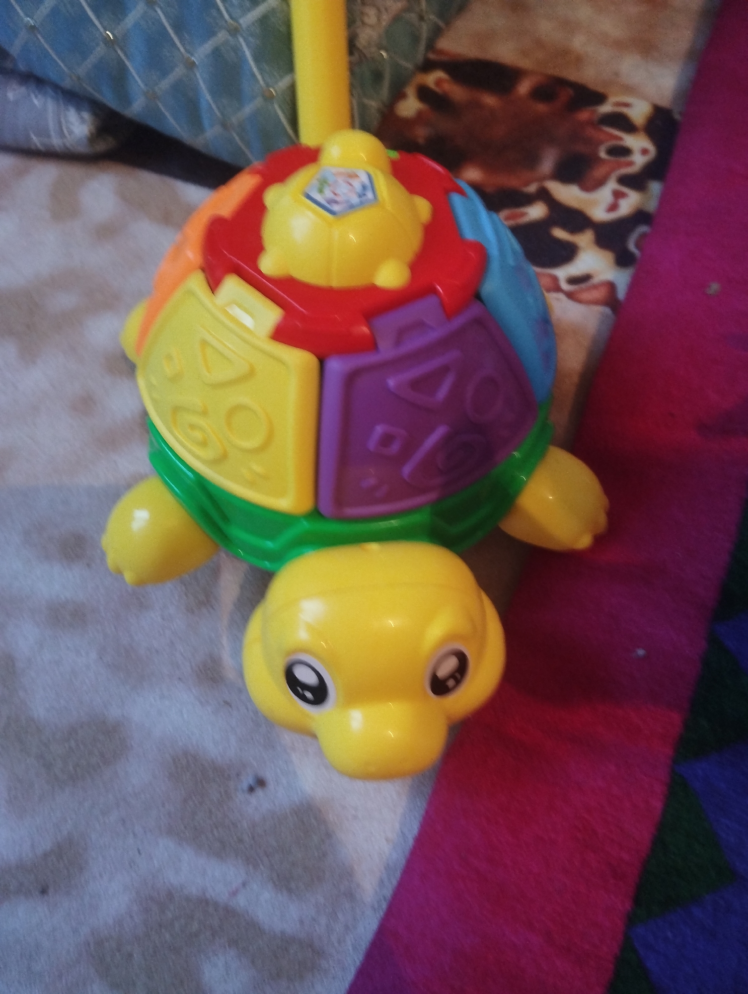 A yellow, plastic toy turtle is lying on its back on a light-colored, patterned rug. The turtle's shell is made of segmented pieces in various colors, including yellow, green, red, purple, and blue. Each segment has a different shape or pattern. The turtle has a yellow head with two large, black and white eyes. The background is out of focus, showing hints of other objects, including what appears to be a blue patterned blanket or fabric in the upper left corner and a pink and purple striped fabric to the right of the turtle. There is no visible indication of the city of Orlovka, Kyrgyzstan, or any environmental details that would suggest this location.