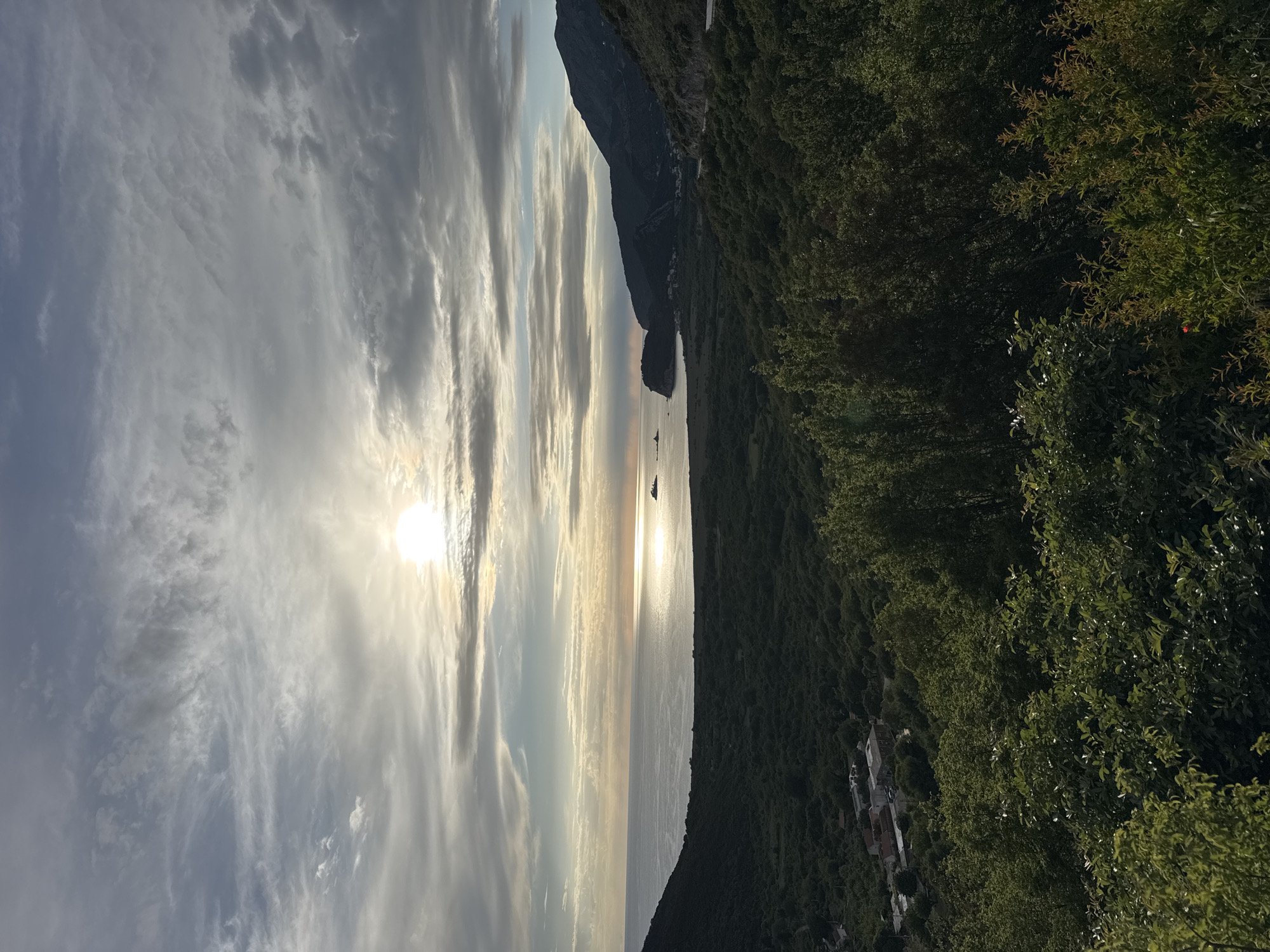 The image depicts a coastal scene, possibly near Kanjoši, Montenegro, at what appears to be sunrise or sunset. The sky is filled with striated clouds, with the sun visible through them. Below the sky is the sea and a portion of coastline. On the right is a tree-covered slope with a few visible buildings. In the sea are two small silhouettes, possibly of boats. A rocky coastline rises up from the sea and is covered in vegetation, as well.