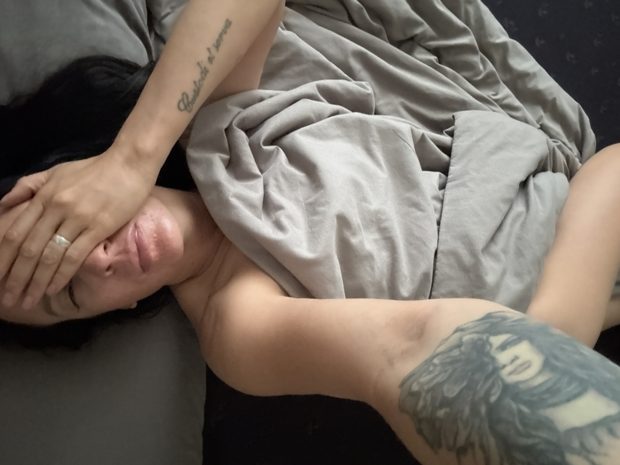 A person is lying in bed, covered by a gray blanket. The person's face is partially obscured by their hand, and they have dark hair. There is a tattoo on their left arm that reads "Custodi et serva" and another on their right upper arm depicting a woman's face. The background is dark. The image does not provide sufficient information to determine its location in the city of Chaikovsky, Russia.