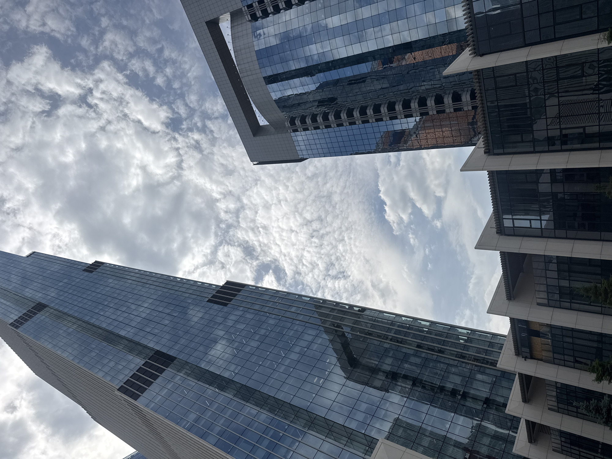 In Moscow, Russia, two modern skyscrapers dominate the frame, viewed from a low angle looking upwards. The skyscraper on the left is significantly taller and sleeker, with a facade of reflective blue glass. Its surface is marked by vertical dark strips, possibly indicating structural elements or lighting. On the right, a second skyscraper features a more angular design with a prominent, large rectangular opening at its top. This building also has a glass exterior, reflecting the sky and the surrounding environment, including a hint of a reddish-brown building. Below these towering structures, a lower, multi-story building with a darker glass facade and distinctive angled white supports is visible. The sky above is filled with a mix of white and grey clouds, suggesting a partly cloudy day. No people are visible in the image.