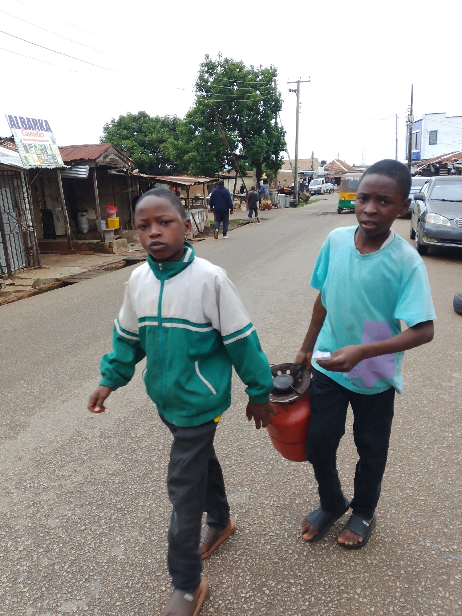 Two young boys are walking on a paved road in what appears to be a market area. The boy in the foreground on the left is wearing a green and white jacket and black pants, and is walking away from the camera. The boy on the right is wearing a light blue t-shirt with a purple design and black pants, and is walking towards the camera while carrying a red propane tank with a stove attachment. In the background, there are several stalls with corrugated metal roofs and signs, including one that reads "ALBARKA Cosmetics." Further down the road, there are other people, vehicles, and buildings, suggesting a bustling urban environment. The sky is overcast.