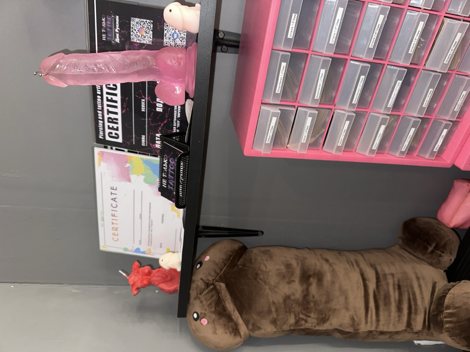 This image displays a section of a professional interior, identified as a piercing and tattoo studio named "Не только tattoo," associated with "Dik Ruslan," located in Saratov, Russia. The walls are a uniform grey.
A black wall-mounted shelf holds several items: On the left, a red candle shaped like a female torso stands next to a small, white, stylized phallic figure. Two framed certificates are positioned centrally; the left one has a colorful abstract background, while the right one, with a black and pink design, explicitly states "Piercing and tattoo artist CERTIFICATE" and includes text in Russian identifying "Не только tattoo" and "Dik Ruslan" with QR codes. A black desk stand displaying "НЕ ТОЛЬКО TATTOO" (Not Only Tattoo) and "ПИРСИНГ" (Piercing) is placed between the certificates. On the right side of the shelf, a large, translucent pink phallic object, appearing to be a dildo, stands upright with a small metal ring attached to its tip, suggesting a piercing demonstration model. Beside it is a smaller, pink, stylized phallic figure.
Below the shelf, a large, brown plush toy, shaped like a cartoonish penis with simplified facial features, leans against the wall. To the right, a pink multi-drawer storage unit is visible, containing numerous clear plastic drawers. Each drawer is labeled in Russian, specifying various types and sizes of piercing jewelry or tools, such as "Тайбон 1.2*5мм" (Tailbone 1.2*5mm), "Микробанелла 1.2*5мм" (Microbanana 1.2*5mm), and "Штанга 1.6*8мм" (Barbell 1.6*8mm). A small pink soft object is partially visible on the floor next to the large plush toy. No individuals are present in the scene, and no actions or interactions are depicted.