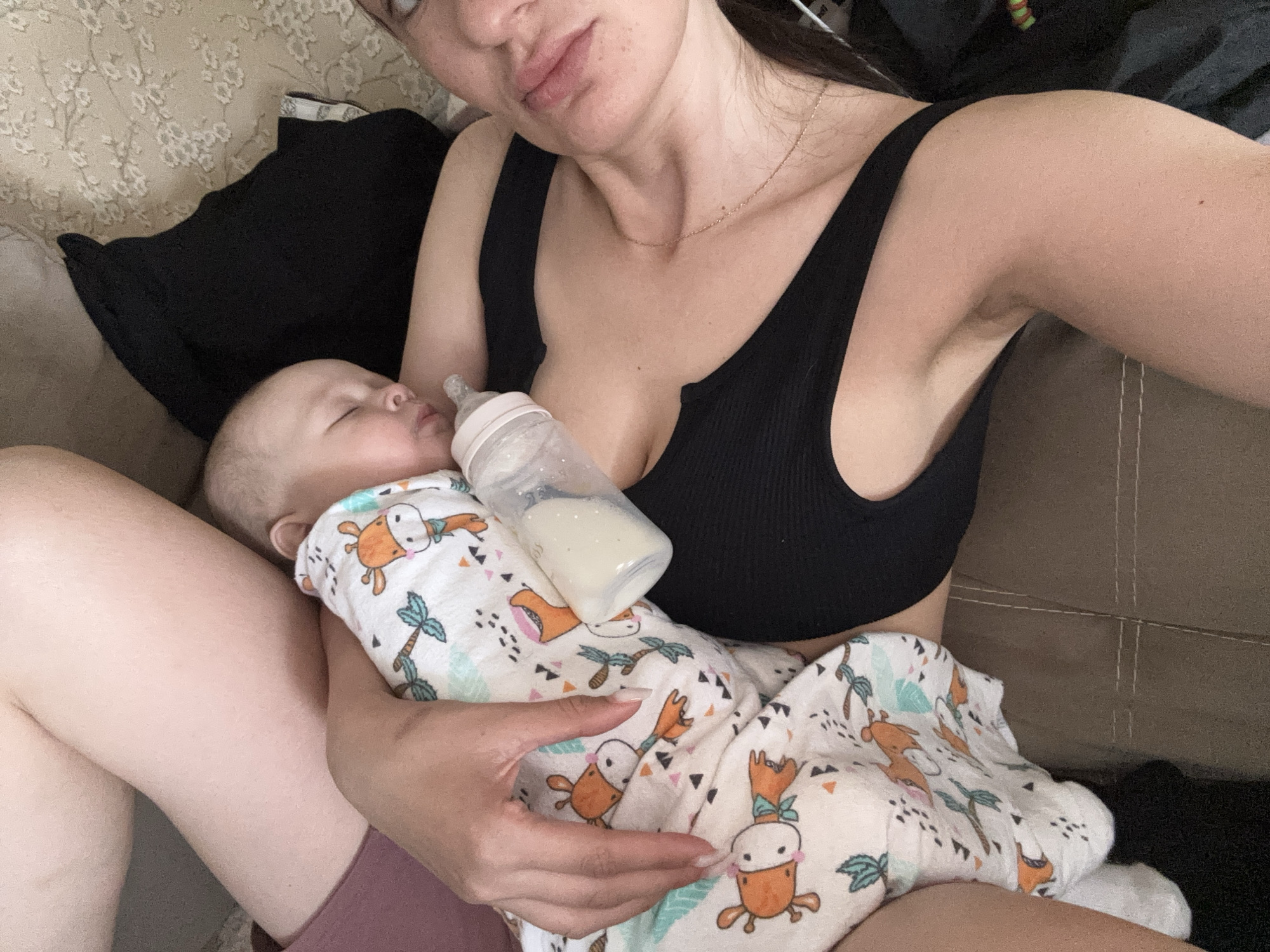 A woman wearing a black tank top is holding a baby who is wrapped in a blanket with a cartoon giraffe and palm tree pattern. The baby is lying down with a bottle in their mouth. The woman is sitting on a brown couch. The location is not discernible from the image.