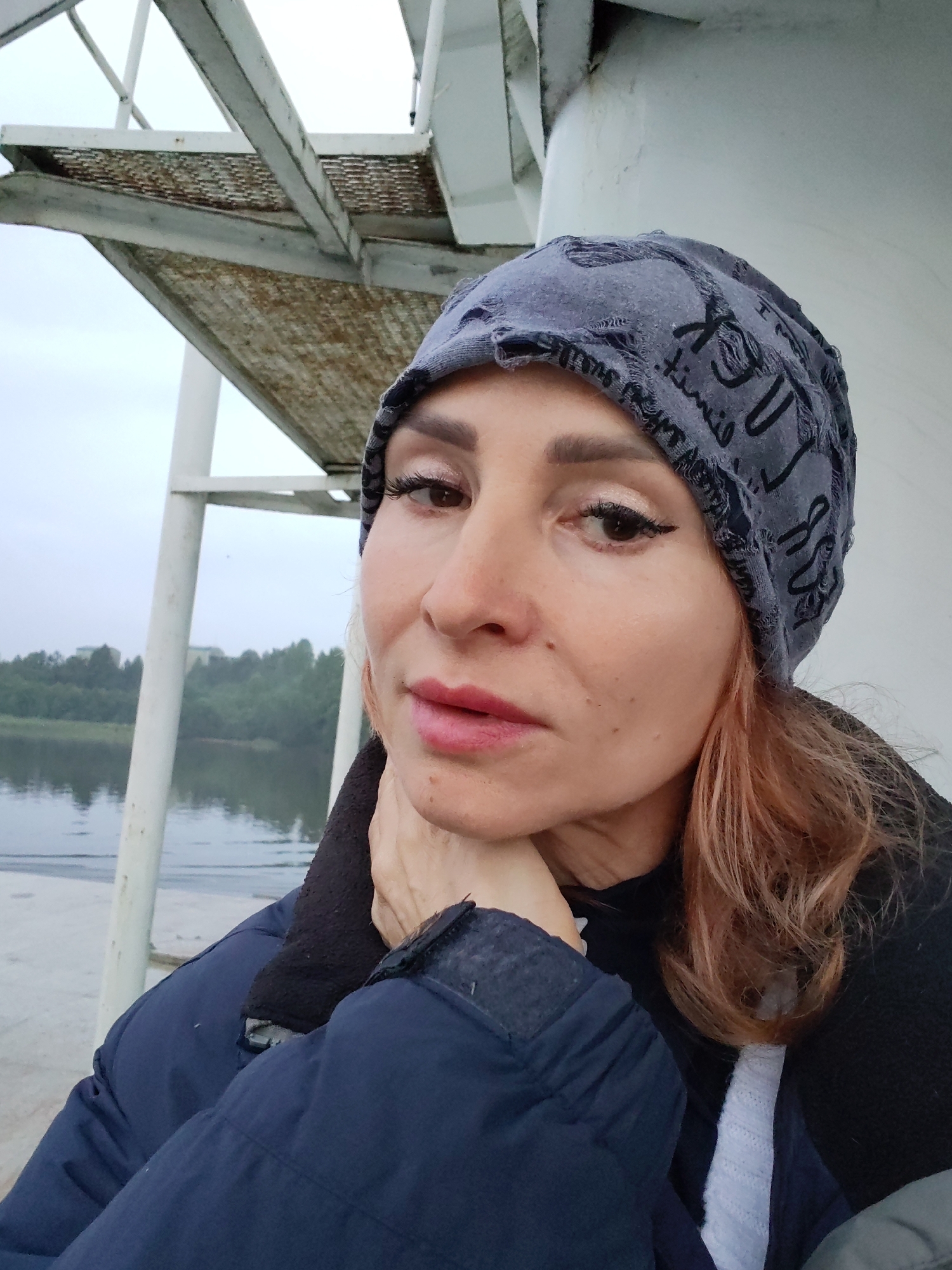 A medium shot captures a woman wearing a grey beanie and a dark blue jacket, positioned in the foreground against a backdrop of a cloudy sky and a body of water with a treeline. The woman is looking towards the camera with her right hand resting on her chin. Her hair, a shade of reddish-brown, is visible on the right side of the frame. In the background, a white structure with metal stairs is visible to the left, and a body of water is seen with trees on the opposite bank. The location is identified as Ivanov Bor, Russia.