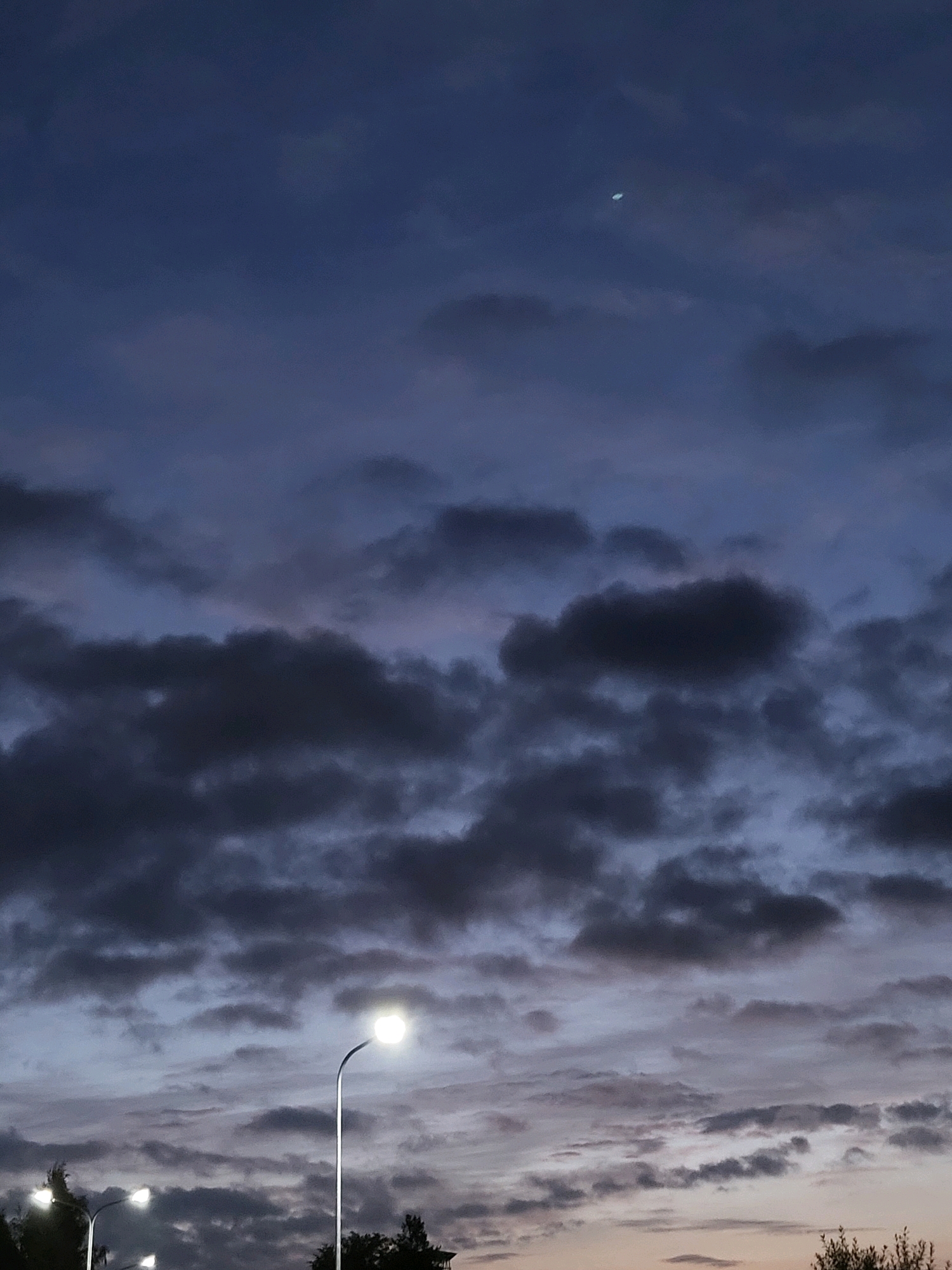 The image depicts a twilight sky over Ozolnieki, Latvia. The sky is filled with a mix of dark and lighter clouds, with a sliver of the moon visible in the upper right quadrant. In the foreground, several streetlights are illuminated, casting a warm glow. One streetlight is prominently featured on the right side of the image, while another is partially visible on the left. Silhouettes of trees and some foliage are visible at the bottom of the frame, suggesting an outdoor, possibly urban or suburban, setting. The overall atmosphere is calm and serene, indicative of the transition from day to night.