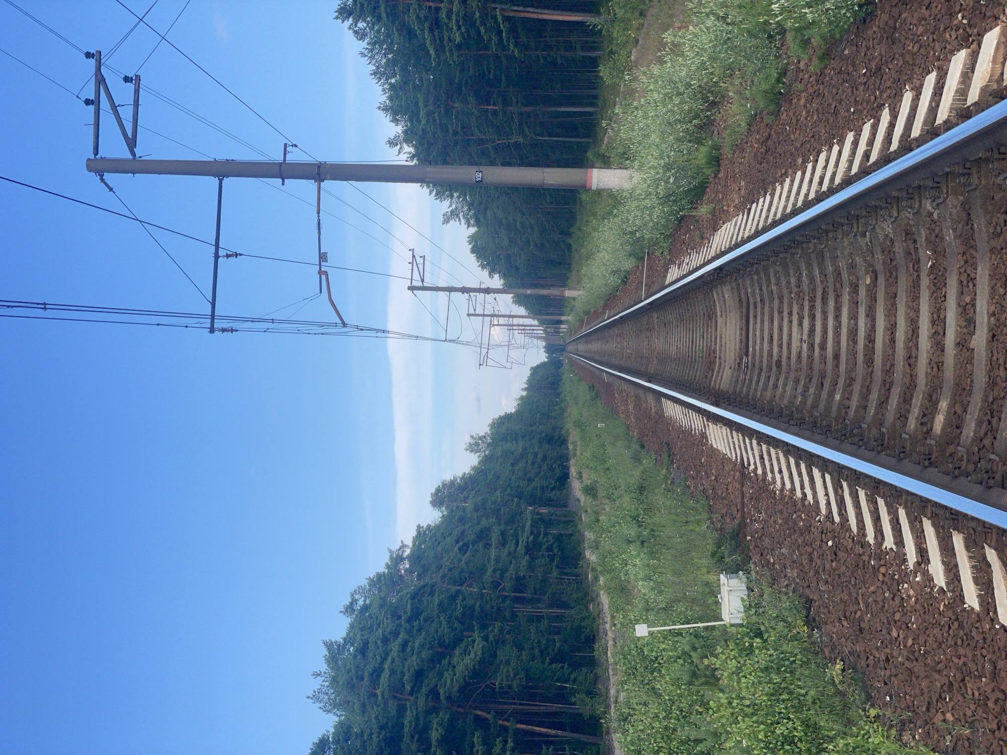 The image depicts a straight railway track receding into the distance, flanked by a dense forest of pine trees on both sides. Overhead, a network of power lines supported by concrete poles stretches along the track. The sky is a clear blue with a few wispy clouds. The railway bed consists of dark brown ballast and concrete sleepers. In the foreground, on the left side of the track, a small white marker post stands next to a light-colored rectangular box. The scene appears to be set in a rural or natural environment, possibly outside of the city of Lebyazhye, Russia, given the typical landscape associated with Russian railways in forested areas. There are no people visible in the image.