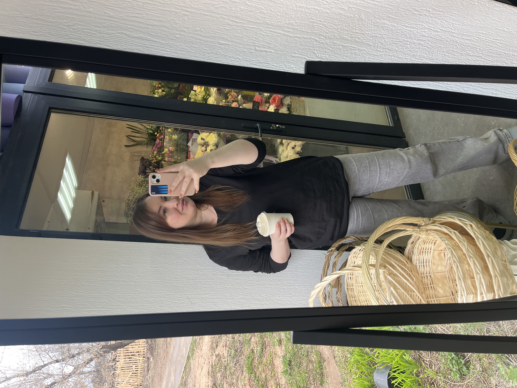 The image shows a woman taking a selfie in a mirror. The mirror is framed in black and reflects a scene both inside and outside. Inside, the reflection shows what appears to be a flower shop display with various flowers and plants. The woman is wearing a black shirt and grey pants, holding a white cylindrical object in one hand and a mobile phone in the other. Baskets are visible on the right side of the reflection. Outside the flower shop, trees and grass can be seen through the window of the shop. It is daytime, based on the light in the reflection. The location is presumed to be in Chaikovsky, Russia, based on the prompt instructions.