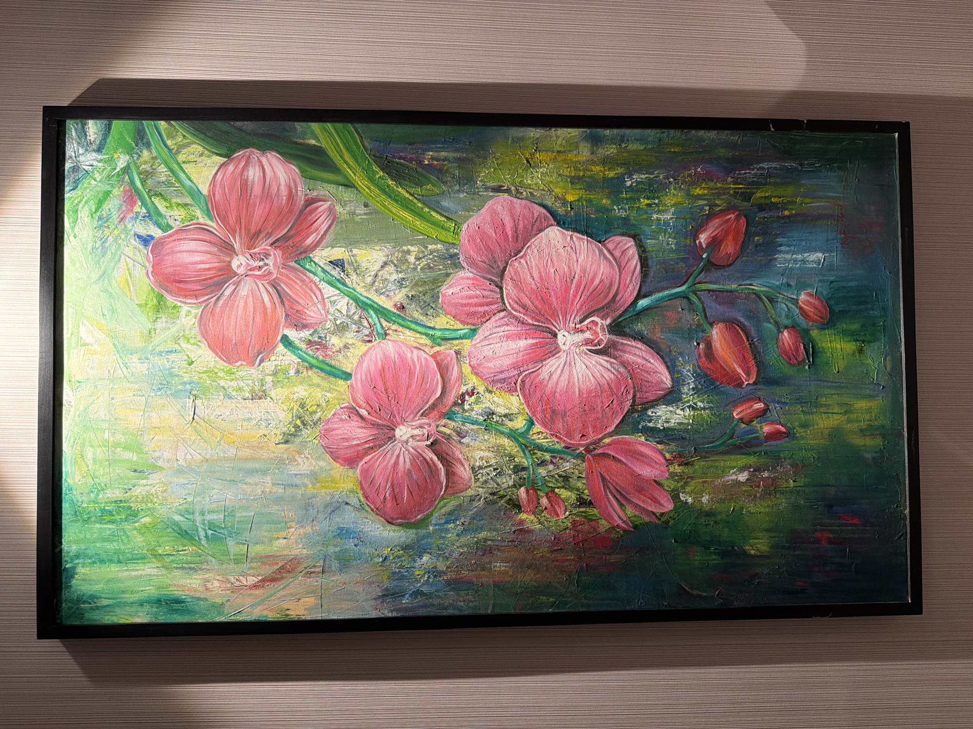An oil or acrylic painting depicting pink orchids is presented in a black frame, hanging on a textured, light brown/beige wall. The painting features several large, vibrant pink orchid blossoms and smaller buds connected by green stems. The flowers exhibit a notable impasto technique, creating a raised, three-dimensional effect with visible brushstrokes.
The background of the painting is abstract and highly textured, with a palette of mixed greens, blues, yellows, and darker tones, suggesting foliage or an impressionistic natural setting. The texture on the background also appears to be achieved through thick paint application.
The painting is well-lit, though unevenly, with a distinct shadow cast by the frame onto the upper left portion of the horizontally striped wall, indicating a light source from the upper left. The frame is simple and black, showing minor wear in places. No activity is taking place within the image, and no visible text is present. The overall impression is a vibrant, textural floral artwork displayed in an indoor setting.