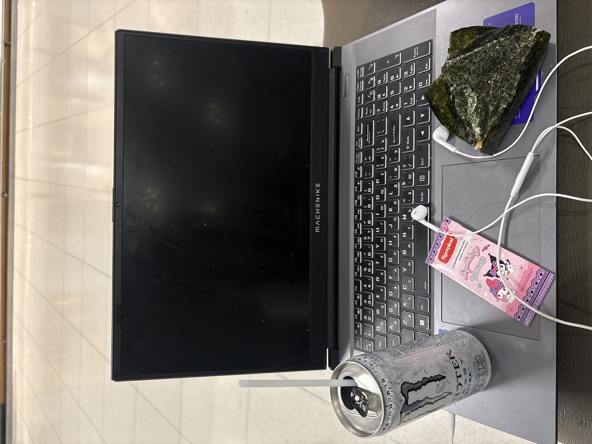 An overhead shot shows a laptop computer with its screen open and facing forward. On the laptop's keyboard, a white can of Monster Energy drink with a straw, a package of Kuromi and My Melody themed bandages, and a piece of onigiri wrapped in nori are visible. A white wired headset is also on the laptop. The laptop is positioned on a desk or table, and the background appears to be an indoor space with a tiled floor and a reflective ceiling. The scene is well-lit, and the objects are in focus.