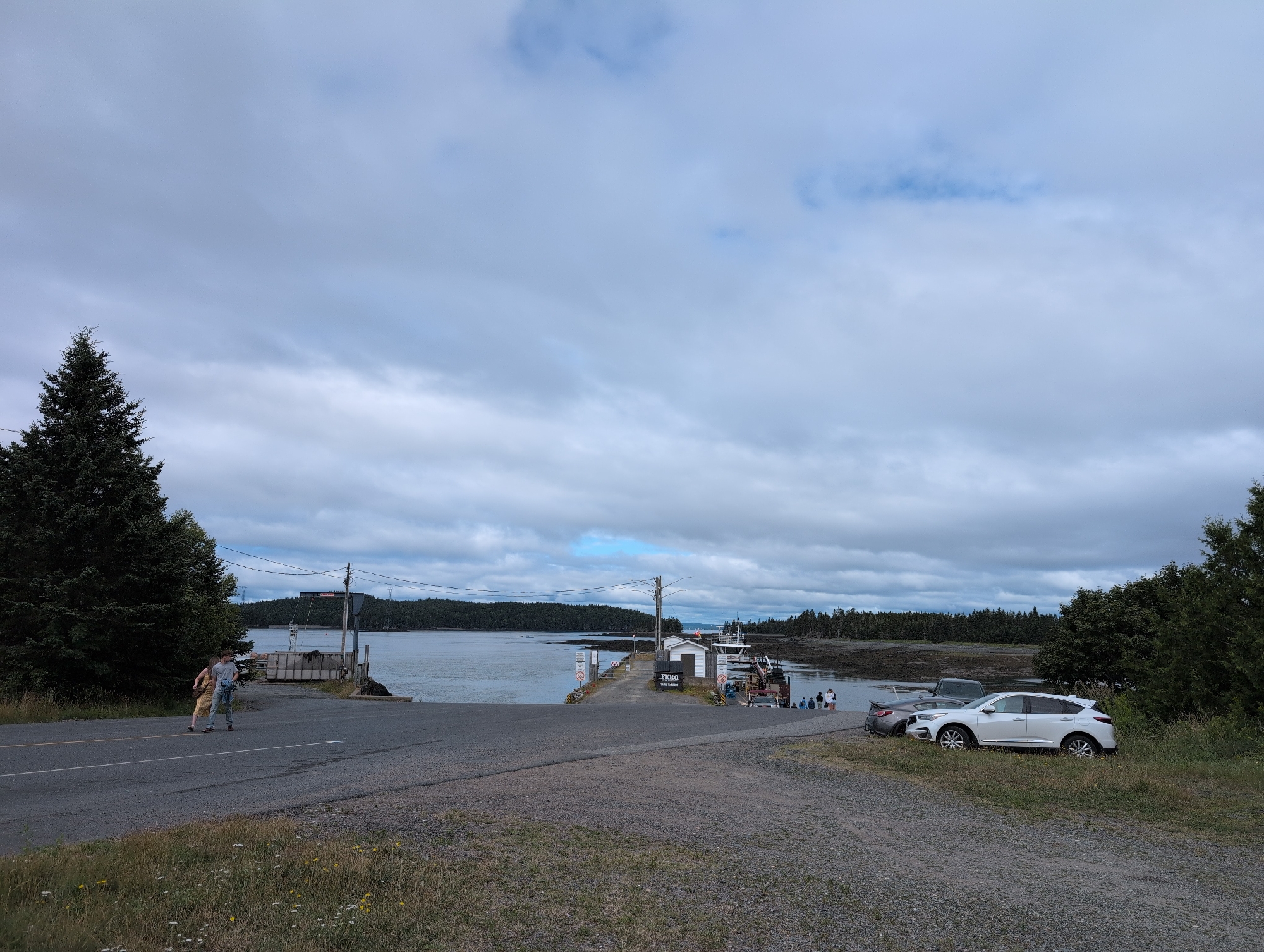 The image depicts a ferry terminal scene in Saint George Parish, Canada, under a cloudy sky. In the foreground, a gravel road curves towards a paved ramp leading down to the water. Two individuals, a person in a yellow dress and another in a gray shirt, are walking on the paved road. To the left, a large evergreen tree stands prominently.
The ferry terminal itself is visible in the mid-ground, featuring a small white building with a sign that reads "FERRY TRANSPORT". A ferry is docked, and several people are gathered on the dock. Beyond the terminal, the water stretches towards a tree-lined shoreline, with a few small islands scattered in the distance. Several cars are parked on the right side of the image, including a white SUV and a dark gray car. The environment is characterized by a mix of paved surfaces, gravel, grass, and vegetation, with the water body and forested shores forming the backdrop.