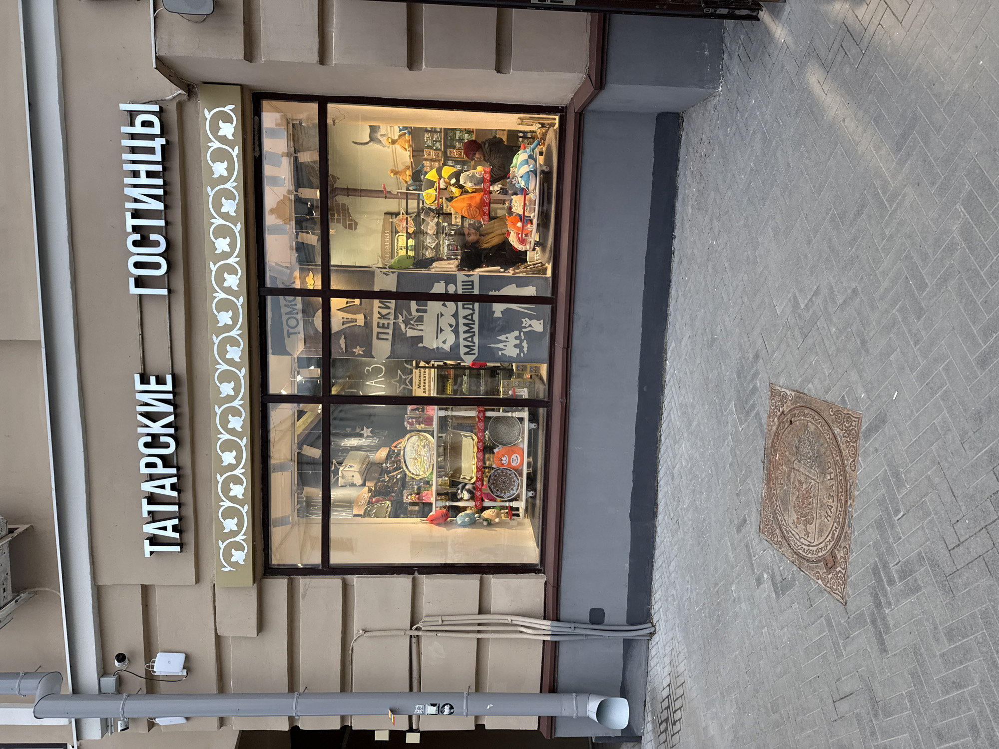 An exterior view shows the illuminated storefront of a shop alongside a gray paved sidewalk. The building facade is light beige with architectural elements. Two vertical signs in white Cyrillic letters are mounted on the facade. The upper sign reads "ГОСТИНИЦЫ" (Gostinitsy) and the lower one "ТАТАРСКИЕ" (Tatarskie), together likely indicating "Tatar Treats" or "Tatar Souvenirs/Gifts." A decorative white panel with floral motifs separates the signs from the shop window.
The large shop window displays various items, brightly lit from within. Visible merchandise includes colorful plush toys (a striped bee, a cat-like figure, other animals), decorative plates, trays, and possibly other souvenirs or traditional goods. Reflections in the window obscure some details, but partial words like "ПЕКИ" (Bake) and "МАМАДЫШ" (Mamadyš – a town in Tatarstan) are discernible on posters or signs inside.
Below the window, utility conduits and a gray downspout are visible. A small surveillance camera and a white rectangular device are mounted on the facade near the "ТАТАРСКИЕ" sign.
The sidewalk is paved with gray herringbone-patterned blocks. Embedded in the sidewalk is a circular, ornate metal manhole cover, set within a square frame. The manhole cover features a crest with a winged dragon/snake and the word "КАЗАНЬ" (Kazan) in Cyrillic, along with "RIVE". The overall lighting suggests either evening or an overcast day, with the shop's interior lights being a primary light source. The scene depicts an urban street in Kazan, Russia.