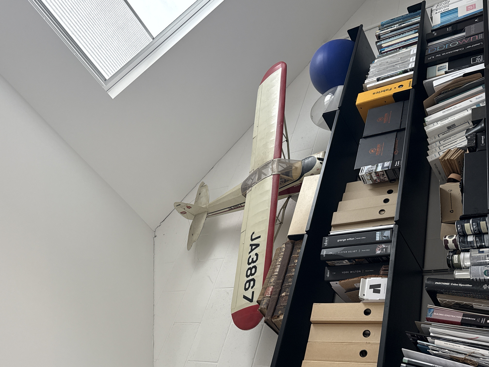 The image depicts an interior space with a window in the upper left and a tall, black bookshelf overflowing with objects on the right. The window is slanted and appears to be in the ceiling, allowing natural light into the room. On the wall, to the left of the bookshelf, is a model airplane with the registration number "JA3867". Next to the bookcase, there is a blue exercise ball and a small clear ball.
The bookshelf is densely packed with various items, including books, binders, boxes, and a few paint cans. The books have titles such as "Fabrics", "PANTONE Color Specifier", "BUILDING CONSTRUCTION HANDBOOK", "BOLON", "grange wilton", "ULSTER VELVET", "YORK WILTON", "Radbo", "marmoleum", and "sphera", suggesting a collection related to design, construction, and materials. The boxes and binders are stacked in an organized manner.
The scene is well-lit, and the composition focuses on the items in the room rather than the specific architecture or location. The clutter and variety of objects suggest a workspace or storage area within a home or office. The image does not provide information about the city of London or the United Kingdom, nor does it describe the presence of any people or interactions within the scene.