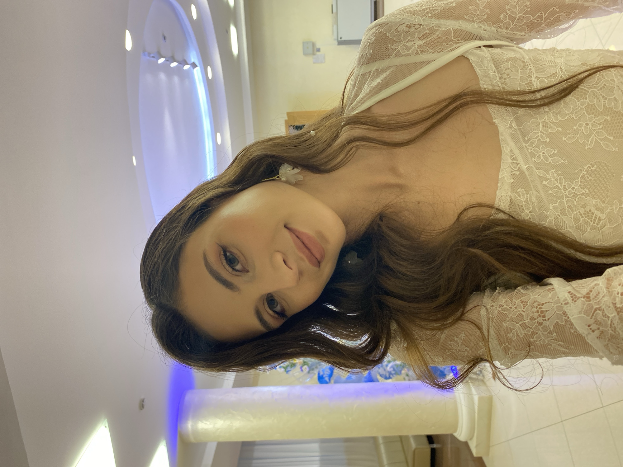 The media file shows a young woman with long, wavy brown hair, wearing a white lace dress. She is posed with her head tilted, looking towards the camera with a gentle smile. A small, delicate flower earring is visible on her ear. The background appears to be an indoor setting with soft, ambient lighting, including some blue accent lighting. There are architectural elements suggesting a modern interior, possibly a room with ornate detailing. The overall mood is elegant and serene. No visible text is present. The location context is Votkinsk, Russia.