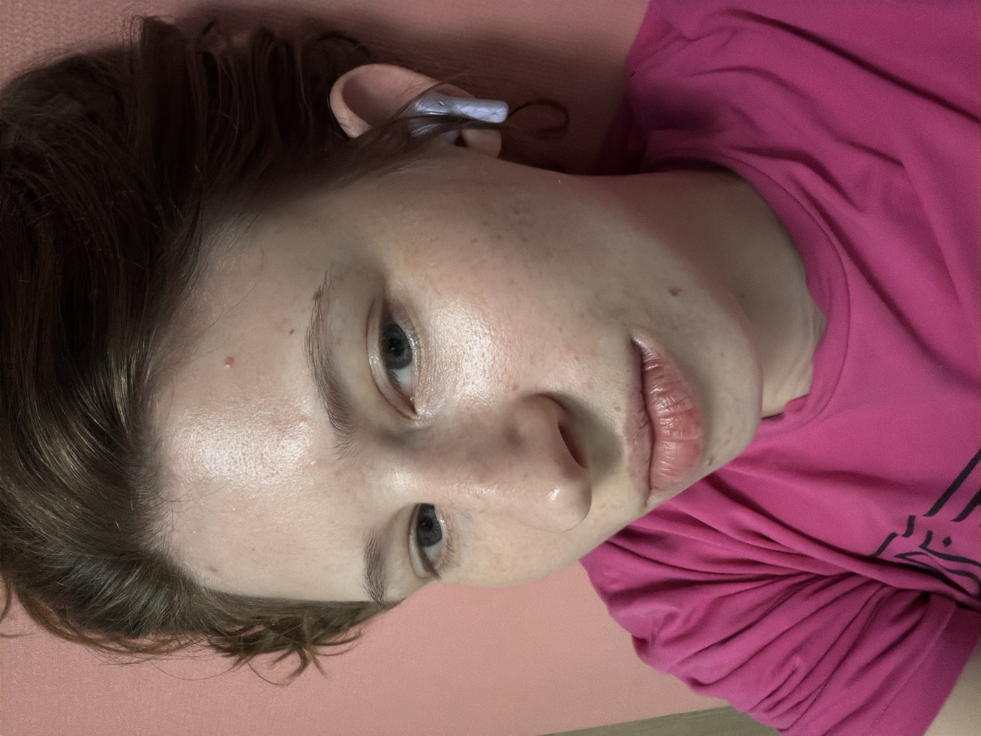 A close-up, overhead shot shows a young person with light skin, freckles, and short brown hair lying down. They are wearing a bright pink t-shirt and have a white wireless earbud in their right ear. The person is looking directly at the camera with a neutral expression. The background is a solid pink surface, possibly a yoga mat. No specific landmarks or details that would indicate the location in Dongducheon-si, South Korea are visible.