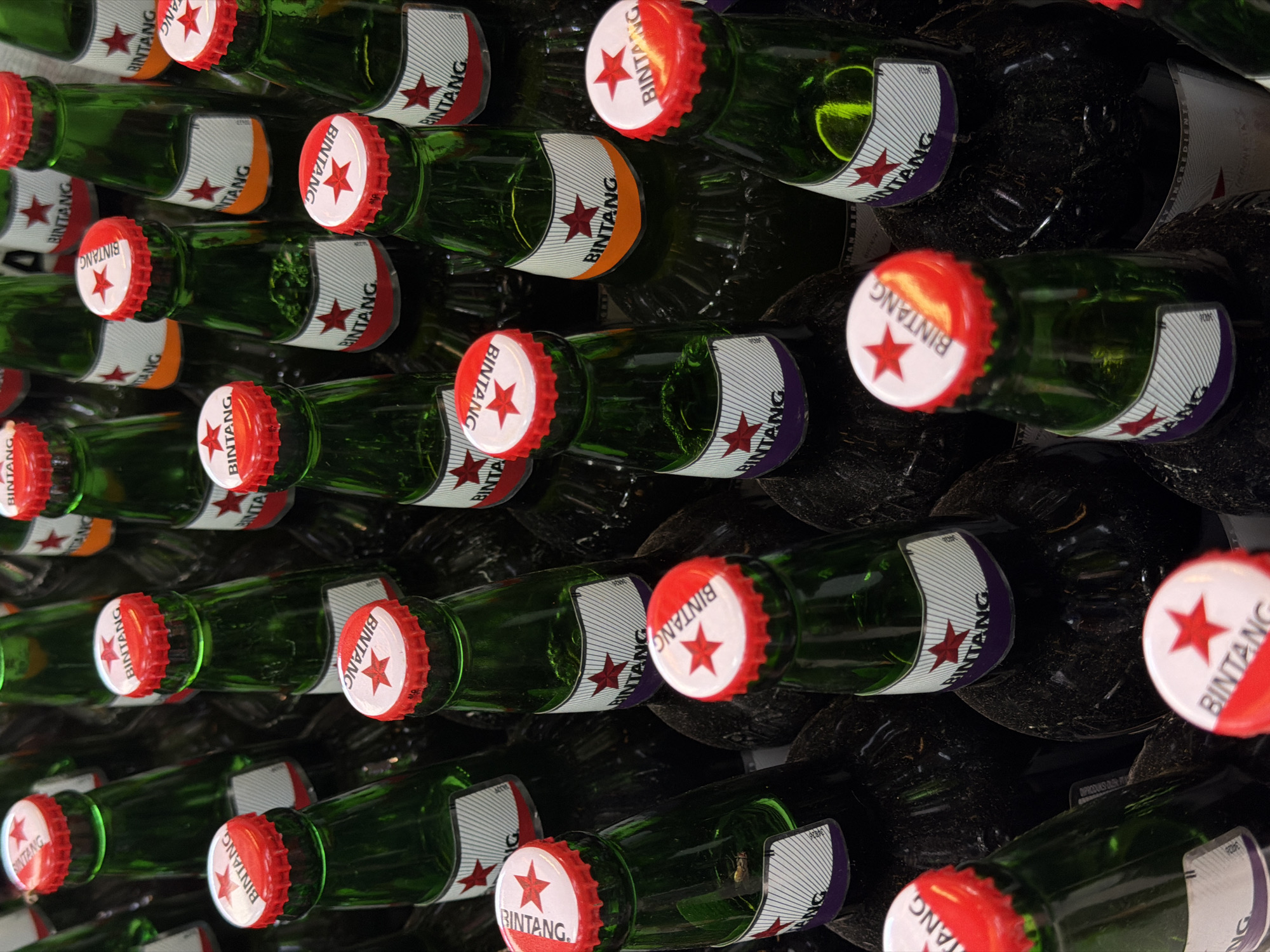 The image shows a close-up of stacked Bintang beer bottles. The bottles are green glass with a white label that reads "Bintang" with a red star. The bottle caps are red and white, also with the "Bintang" logo. The bottles are densely packed and stacked, creating a wall-like effect. Some bottles have different colored labels like orange and purple. The location is North Kuta, Indonesia.