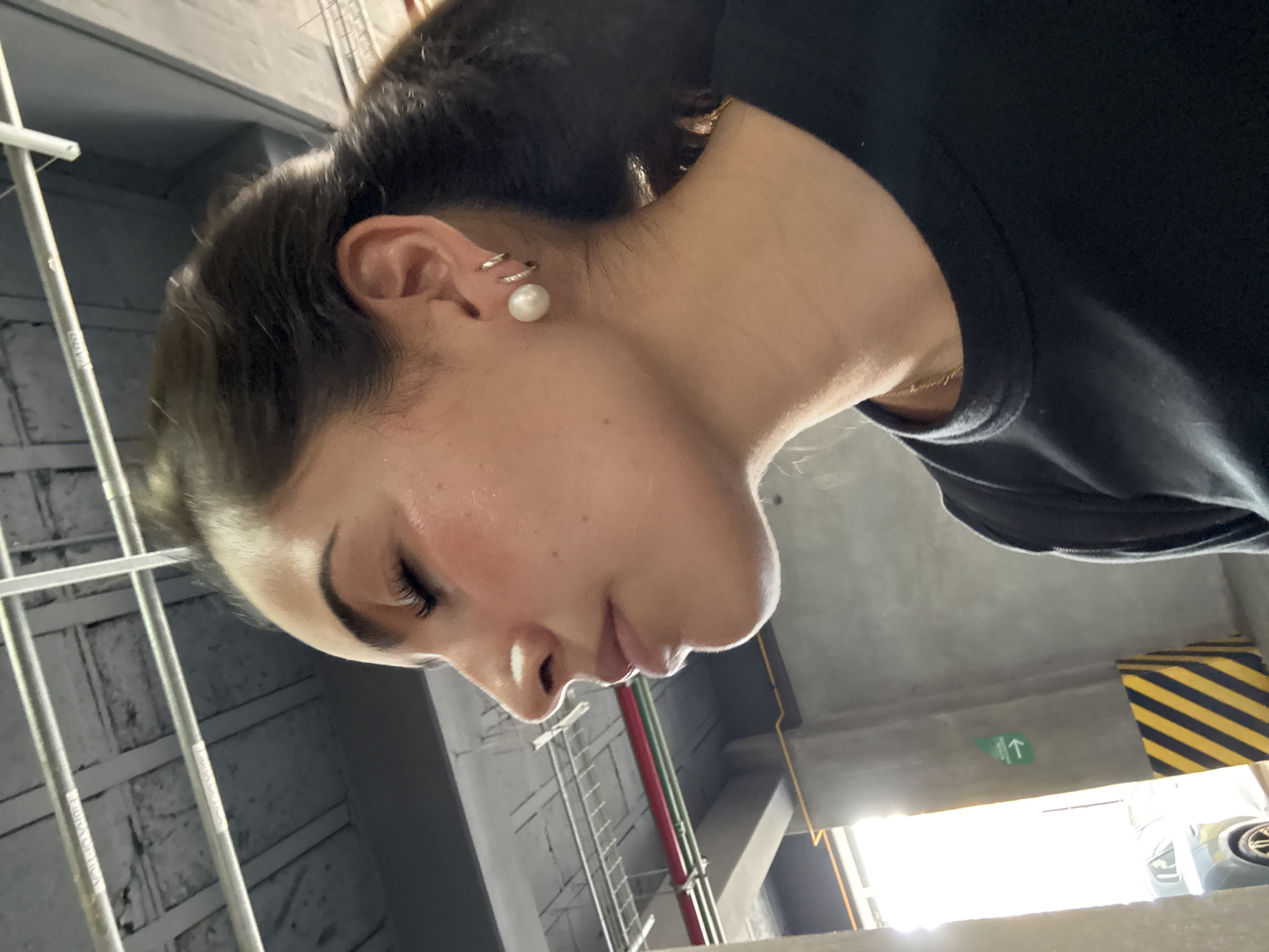 A woman with dark hair pulled back is facing downward and to the left in an underground parking garage in San Bernardino Tlaxcalancingo, Mexico. She is wearing a black shirt and has a gold chain around her neck. The woman has fair skin, dark eyebrows, and long eyelashes. She is wearing earrings, including a pearl stud. The parking garage has visible pipes, beams, and a green emergency exit sign. A section of the wall is painted with yellow and black stripes. There is a car parked in the distance.