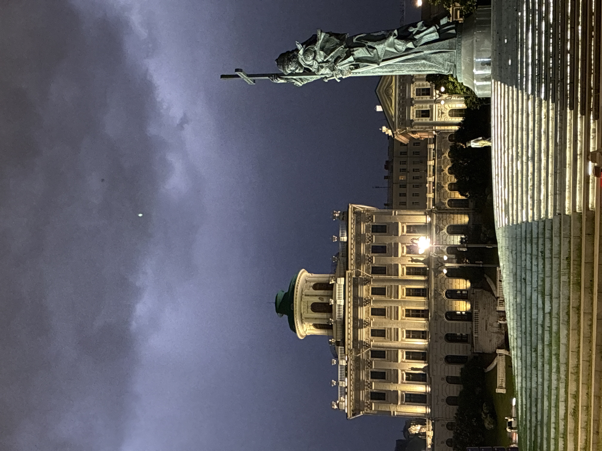 The image shows the Monument to Saint Vladimir in Moscow, Russia, at night. The statue of Saint Vladimir holding a cross is visible at the top right of the image. Below the statue is a building with lit windows. In the foreground are the stairs leading up to the monument. The sky is dark and cloudy.