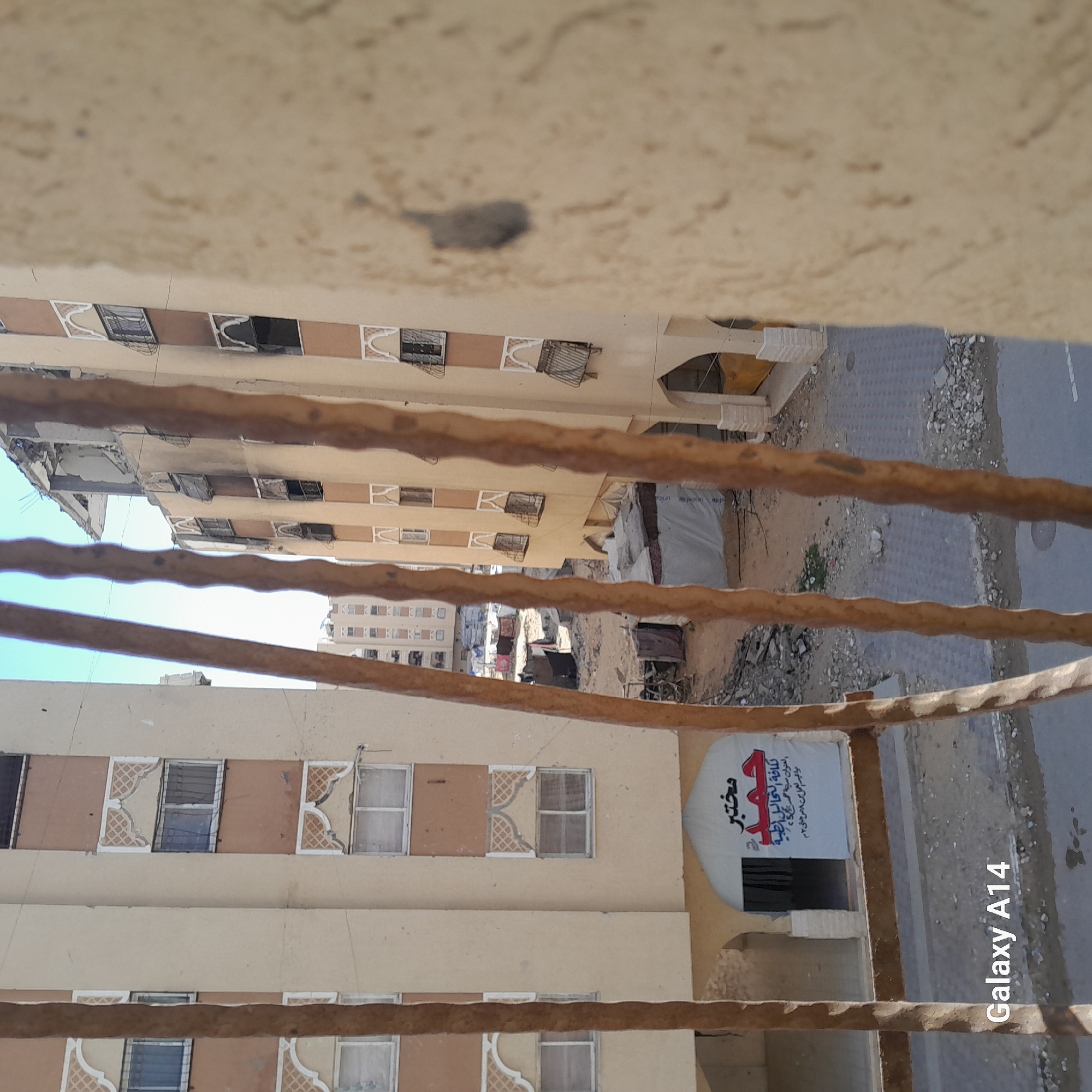 The image displays an urban scene in Khan Yunis, Palestinian Territory, viewed through four rusty, wavy vertical bars in the immediate foreground. To the left, a multi-story building with a light beige facade and reddish-brown sections around windows features decorative white arched frames above each window. A large arched entrance on the ground floor is covered by a white banner displaying Arabic text, including "مختبر" (Laboratory) and "مختبر حمد للتحاليل الطبية" (Hamad Medical Laboratory) in red and black. To the right, another multi-story building of similar architectural style exhibits significant damage; its upper stories are partially collapsed with exposed structural elements and debris, and multiple windows are broken, missing panes, or have bent grilles.
Between these two buildings lies an open, unpaved area covered in dirt and rubble. Within this area, several makeshift shelters or tents are visible, constructed from various materials, including a white tarpaulin featuring a blue "unicef" logo. The ground level in the foreground consists of a paved street with scattered debris. A clear, light blue sky is visible between and above the structures. No people are present in the scene. The bottom left corner of the image contains a watermark "Galaxy A14".