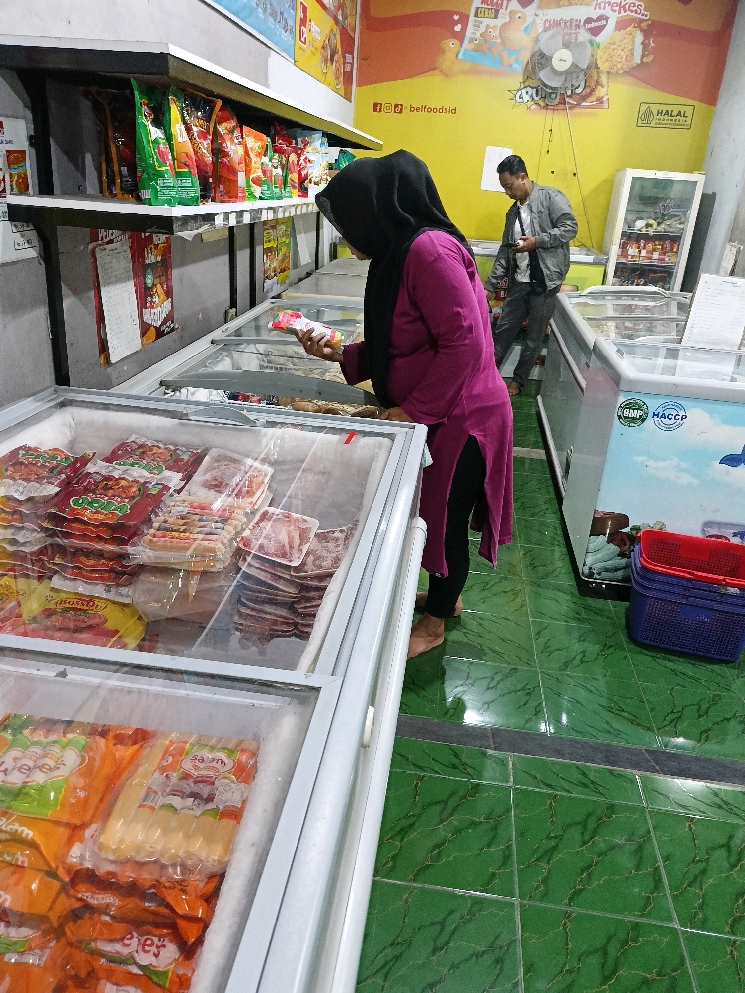 In Manisrenggo, Indonesia, a grocery store interior is captured. Two individuals are present. In the foreground, a woman wearing a black hijab and a magenta tunic is standing, holding a packaged food item and looking into a chest freezer filled with various types of packaged meat products, including sausages. To her right, a man in a grey jacket and dark pants stands, looking at his mobile phone. Behind them, a white display refrigerator and shelves stocked with packaged goods are visible. The floor is tiled in green with a marbled pattern.