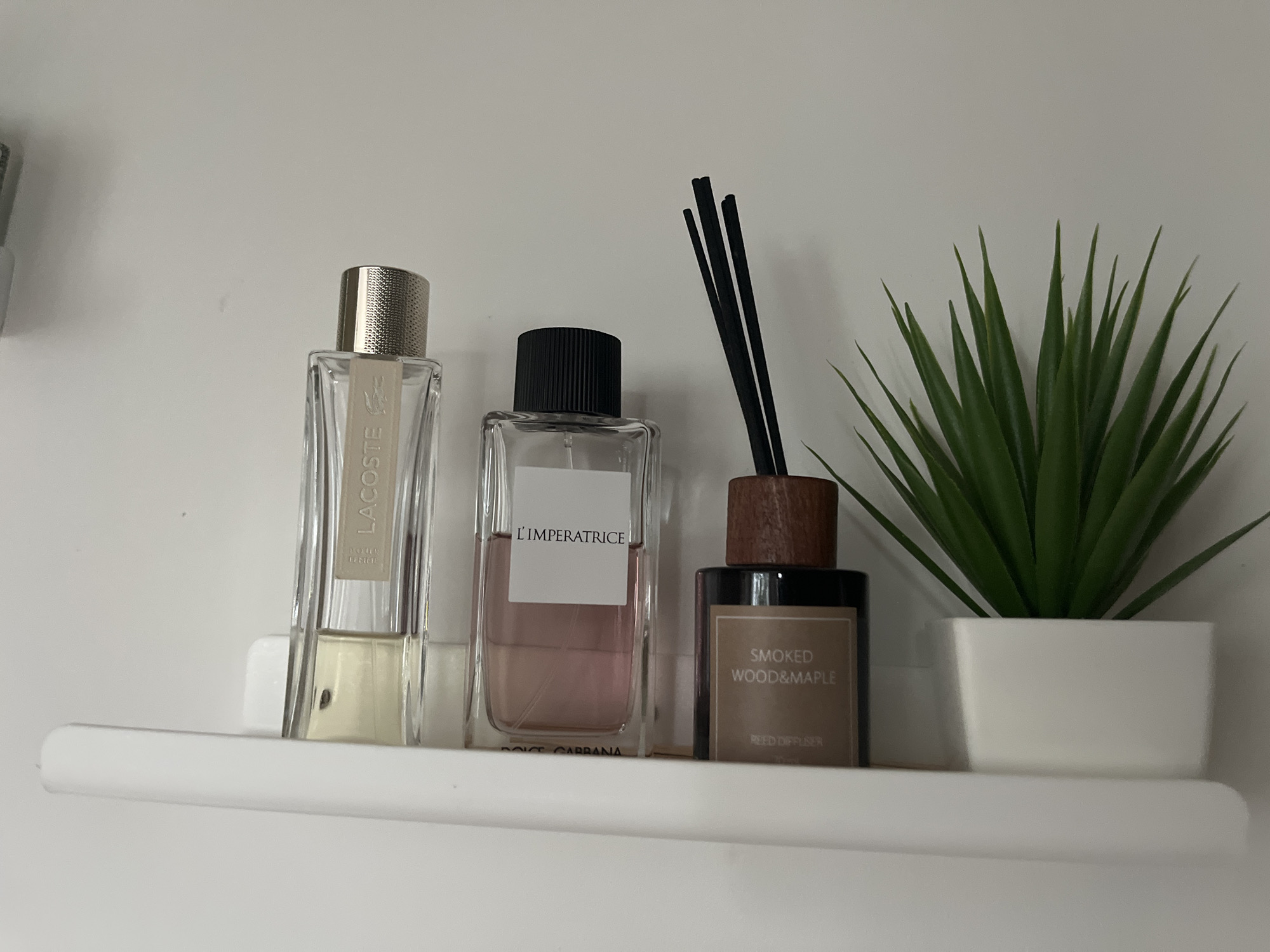 A white shelf displays three decorative items: a clear glass bottle of "LACOSTE" perfume, a clear glass bottle of "L'IMPERATRICE" perfume with pink liquid, and a dark glass reed diffuser labeled "SMOKED WOOD & MAPLE". A small artificial green plant in a white pot is to the right of the reed diffuser. The background is a plain white wall. The scene is likely an indoor setting, possibly a bathroom or bedroom. There is no visible indication of Kohtla-Järve linn, Estonia.