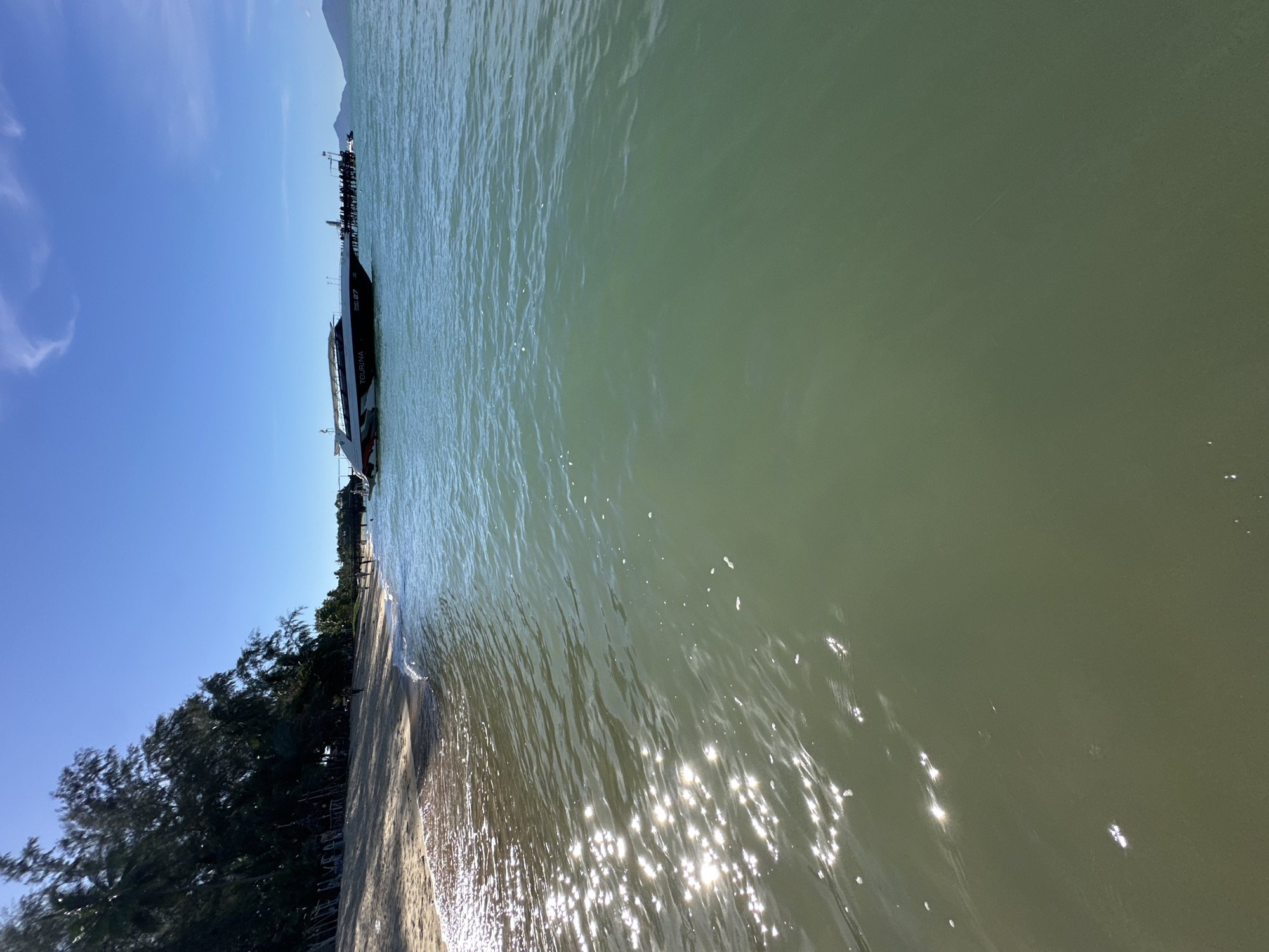 The image depicts a scenic view of a body of water, likely the sea, under a clear blue sky. A ferry, named "TOURISMA 27," is docked near a pier, its dark hull contrasting with the light green water. The ferry appears to be a passenger vessel, suggesting it's either arriving or departing. To the left of the ferry, a sandy beach lined with lush green trees can be seen, indicating a tropical or coastal setting. The sun is shining brightly, creating sparkling reflections on the water's surface, implying it's daytime. The overall atmosphere is tranquil and inviting, characteristic of a beach destination. The name "TOURISMA 27" is clearly visible on the side of the ferry, along with some smaller text that is not legible. The location is identified as Baan Tai, Thailand.