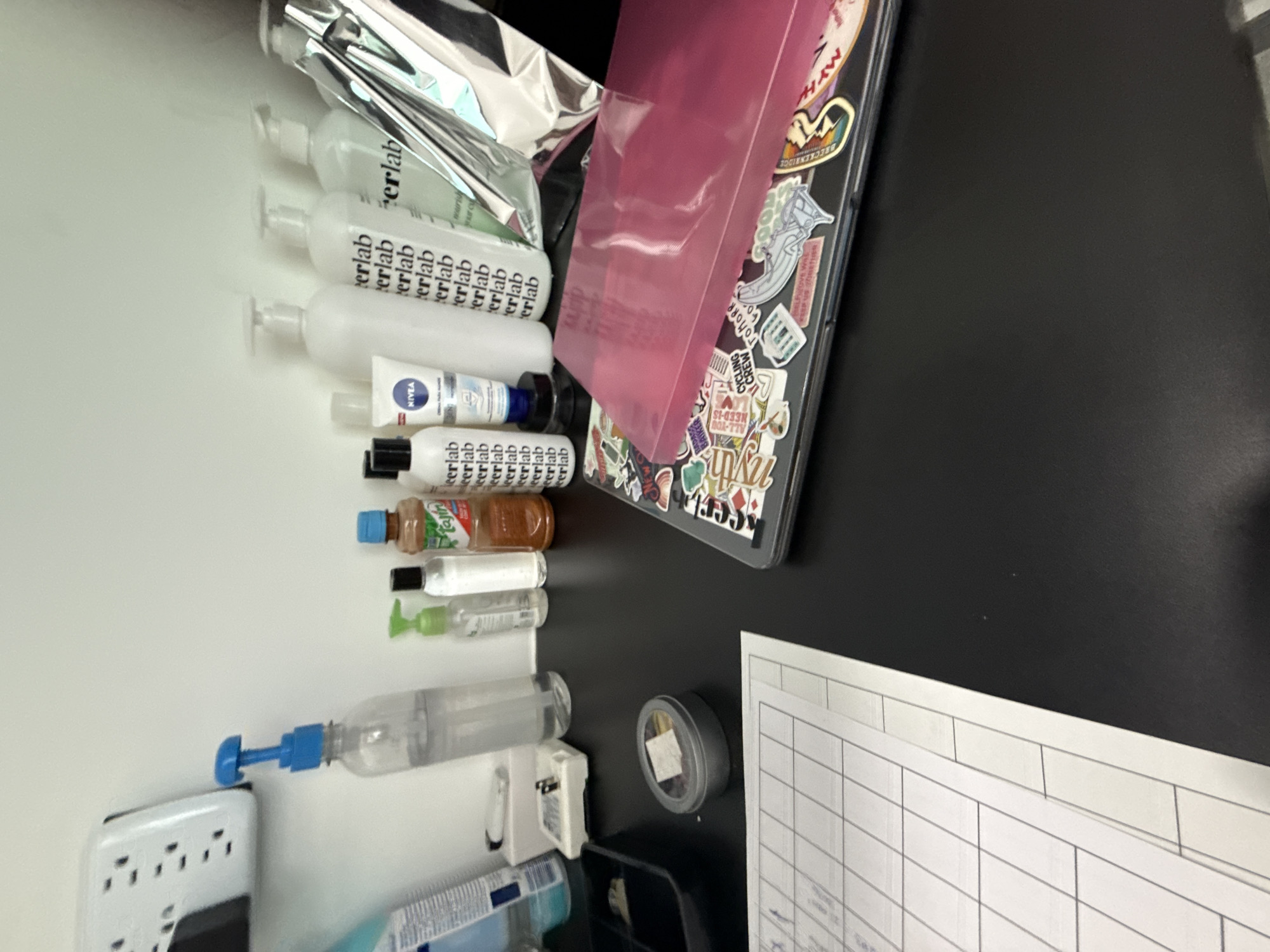 A high-angle shot depicts a cluttered surface with various items. On the left side, there is a row of assorted bottles and tubes, including brands like "erlab" and "NIVEA," ending with a bottle with a blue pump. Next to these, there is a white power outlet with multiple slots. Above the bottles, metallic tubes and a pink box sit atop a laptop decorated with stickers. To the right, there's a round container with a metal lid, followed by a sheet of paper with ruled lines, possibly a form or schedule. The background is divided between a white surface on the left and a black one on the right. The location of San Bernardino Tlaxcalancingo, Mexico, cannot be determined from the visual information provided.