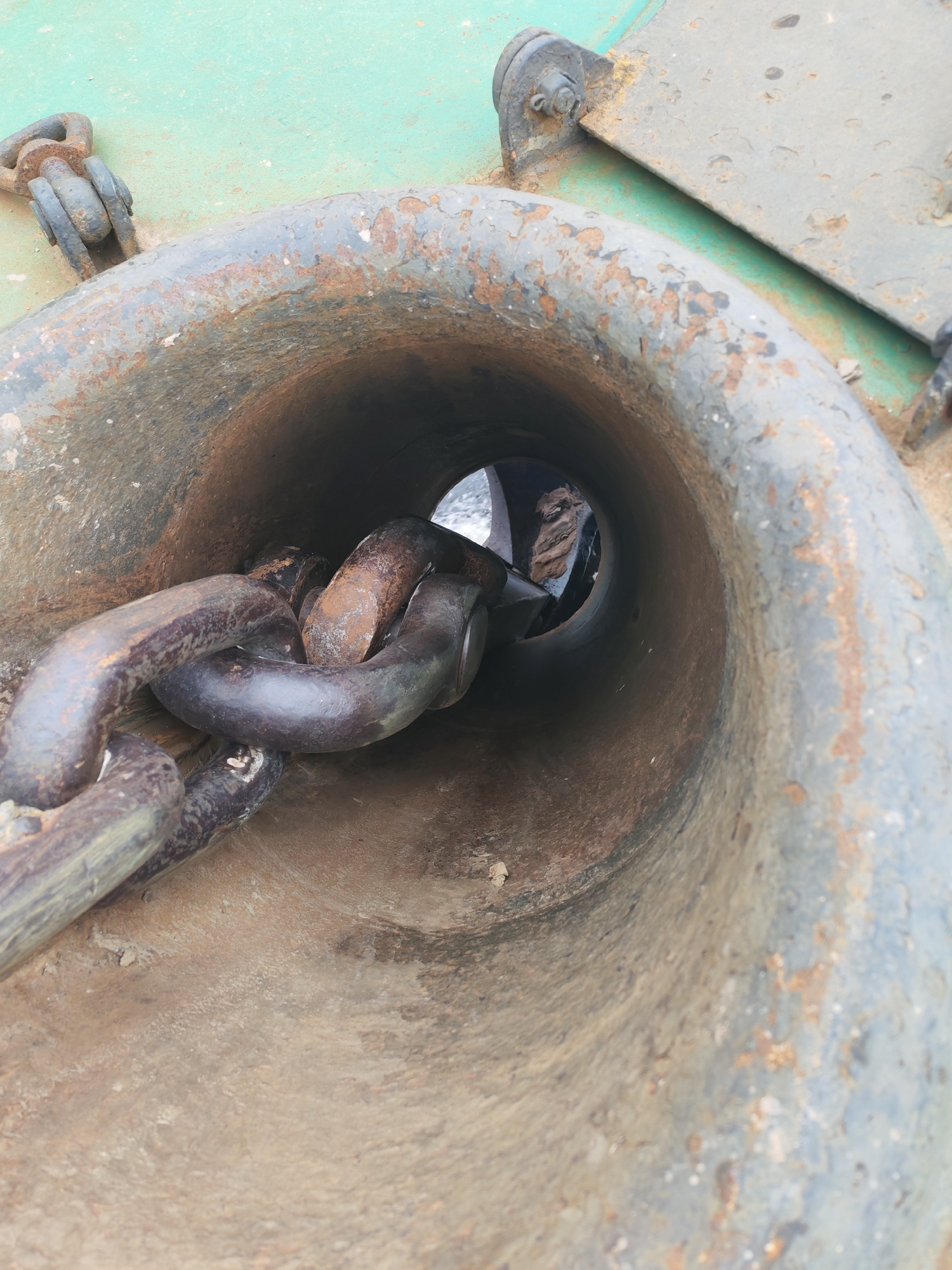 The image captures a close-up, downward perspective of a heavy-duty maritime feature, likely on a ship or in a port setting in Городище, Russia. The central subject is a large, conical metal hawsepipe or chocks, through which a robust anchor chain passes.
The hawsepipe itself is made of dark, weathered metal, showing significant signs of age and corrosion. Its interior and exterior surfaces are covered in pitted rust and a faded, chipped grey-blue paint. The anchor chain, composed of thick, dark grey links, is heavily rusted, particularly in its recesses and where the paint has worn away. Several links are visible, coiling into the hawsepipe and leading into the unseen depths.
Surrounding the hawsepipe is a flat, metal surface painted a muted, faded green. This surface also exhibits considerable wear, with patches of rust and discolored areas, indicating prolonged exposure to weather and salt. In the upper right of the frame, parts of the ship's structure are visible, including bolted fasteners that secure a metal plate.
Through the opening at the end of the hawsepipe, a distant and obscured background is visible, suggesting an open environment, possibly water or a dock. The scene is brightly lit, indicating daytime. The textures are predominantly rough and corroded, highlighting the industrial nature of the equipment and the harsh conditions it endures. The image portrays a static moment; no direct activity is taking place. There is no visible text.